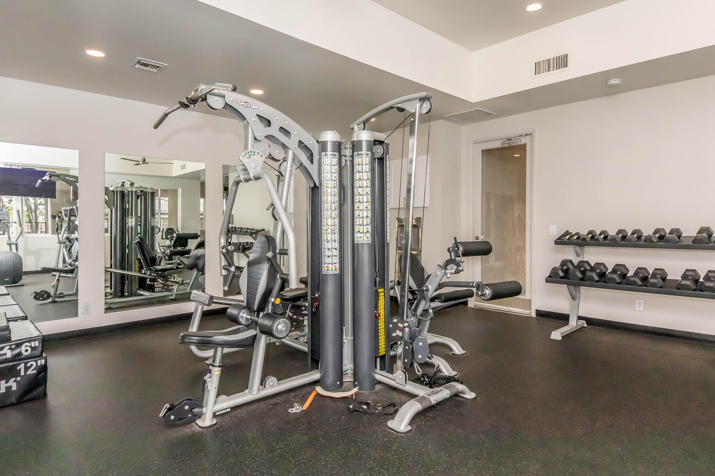 A modern gym interior featuring a multi-station workout machine in the center, surrounded by various exercise equipment. There are mirrors on the wall and a rack of dumbbells in the background, with a clean, well-lit environment conducive to fitness activities.