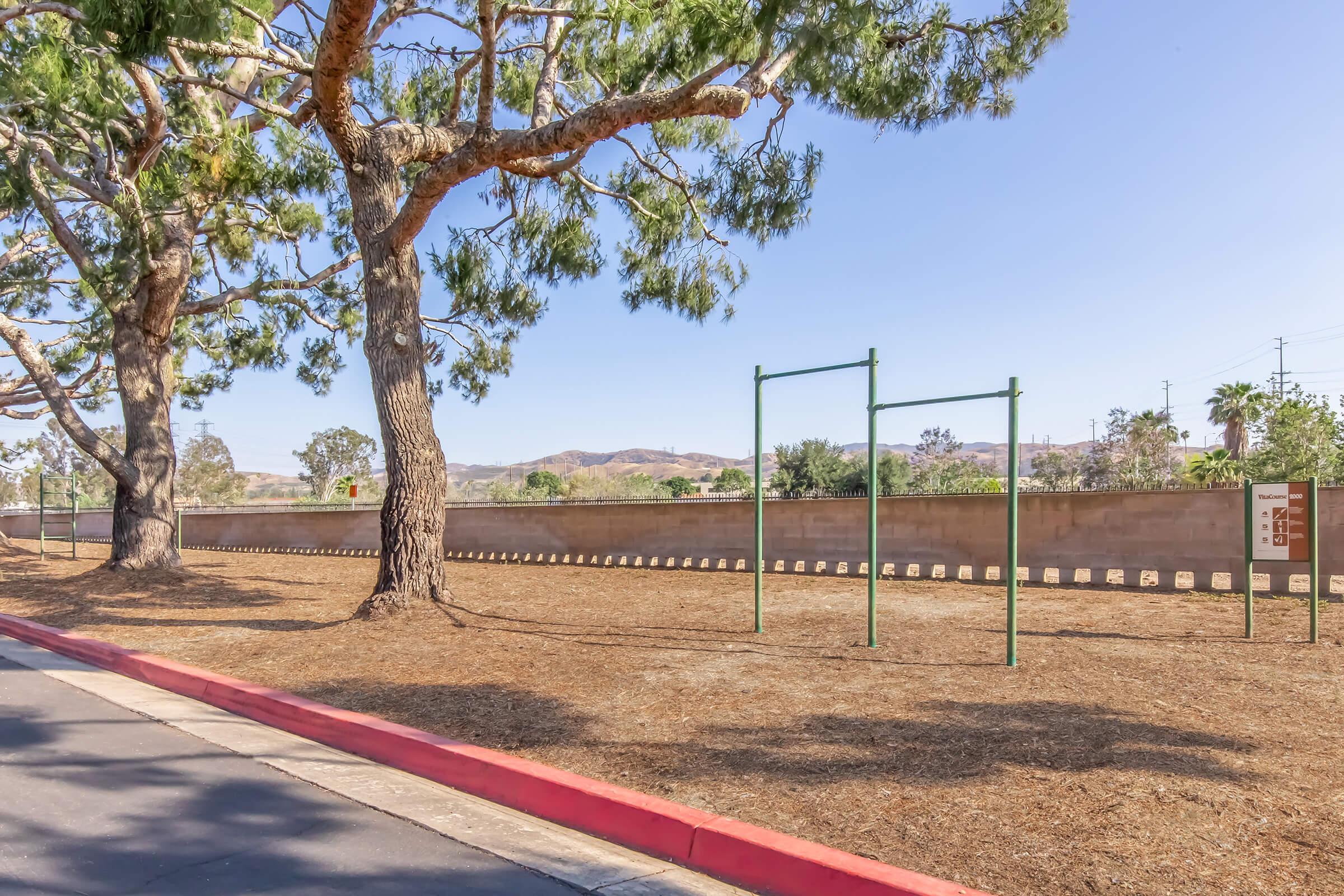 A spacious outdoor area featuring a grassy landscape with a large tree on the left. There are playground pull-up bars positioned on the right side, with a low wall in the background. The scene is set against clear blue skies and distant hills, creating a tranquil and inviting environment.