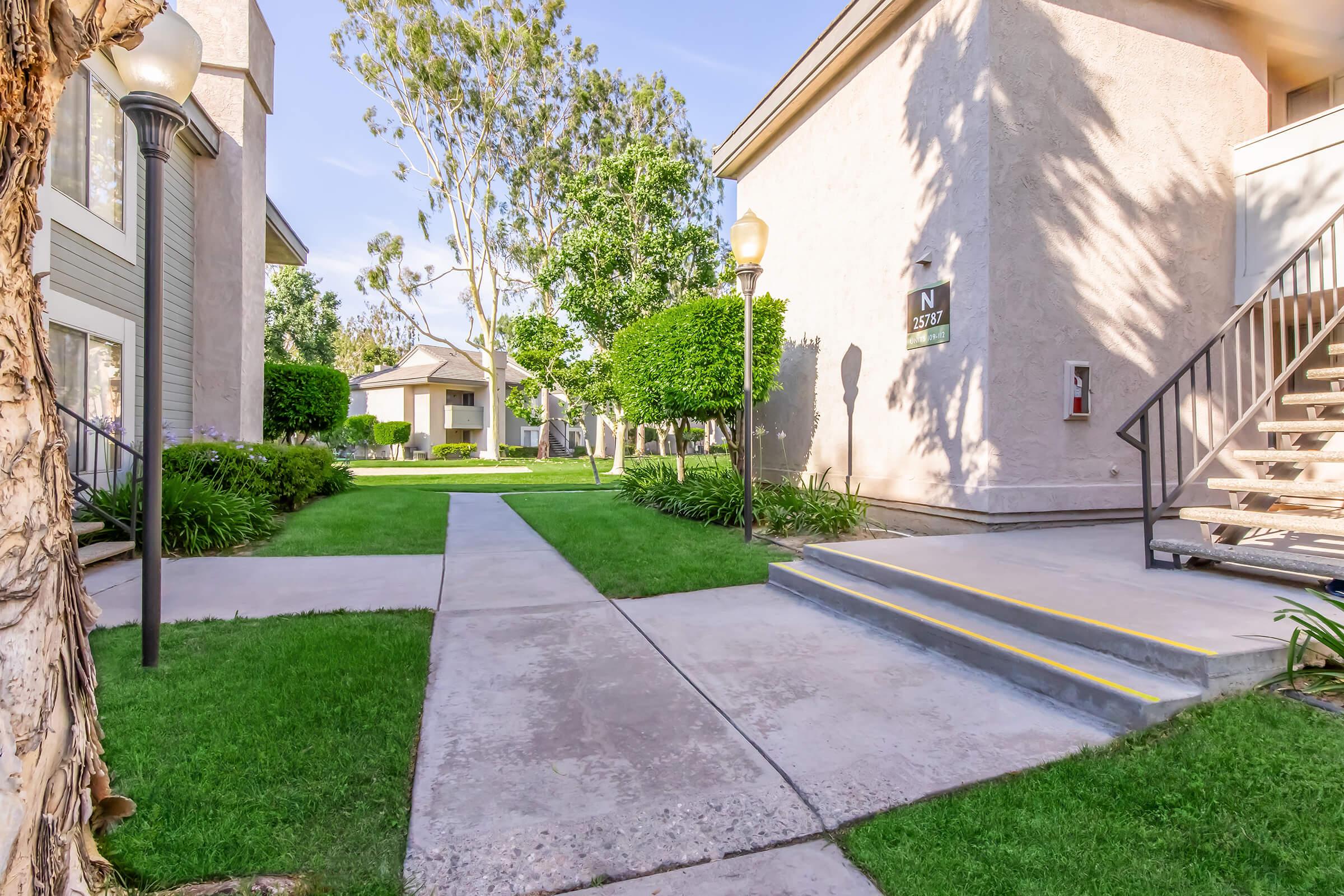 View of a well-maintained apartment complex featuring landscaped gardens, neatly trimmed bushes, and a walkway leading to the entrance. There are staircases and light posts lined along the path, creating a welcoming atmosphere in a sunny, green environment.