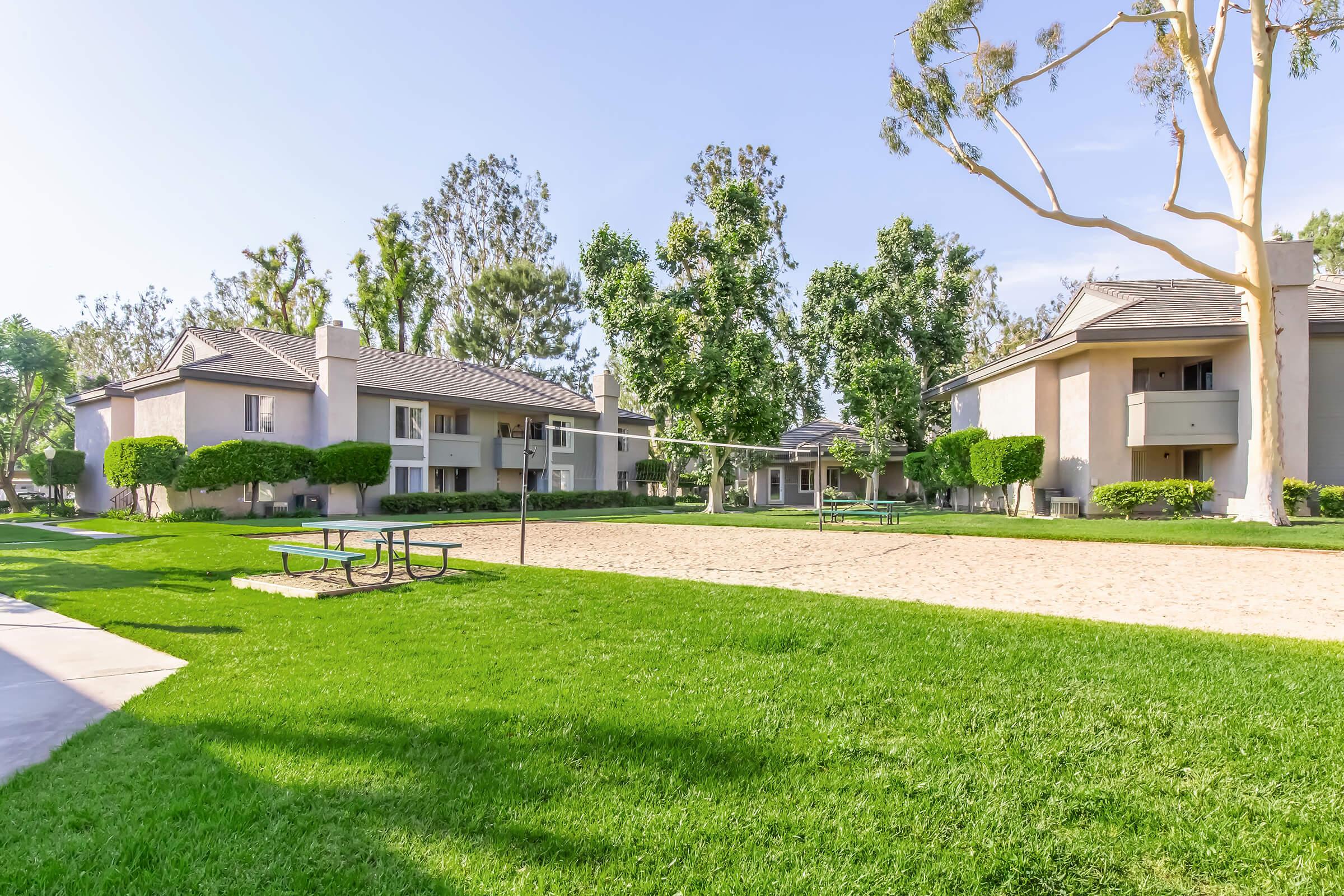 A sunny outdoor area featuring a volleyball court surrounded by well-maintained grass. There are several apartment buildings in the background, along with neatly landscaped trees and bushes. A picnic table is placed nearby, offering a relaxing space in the community.