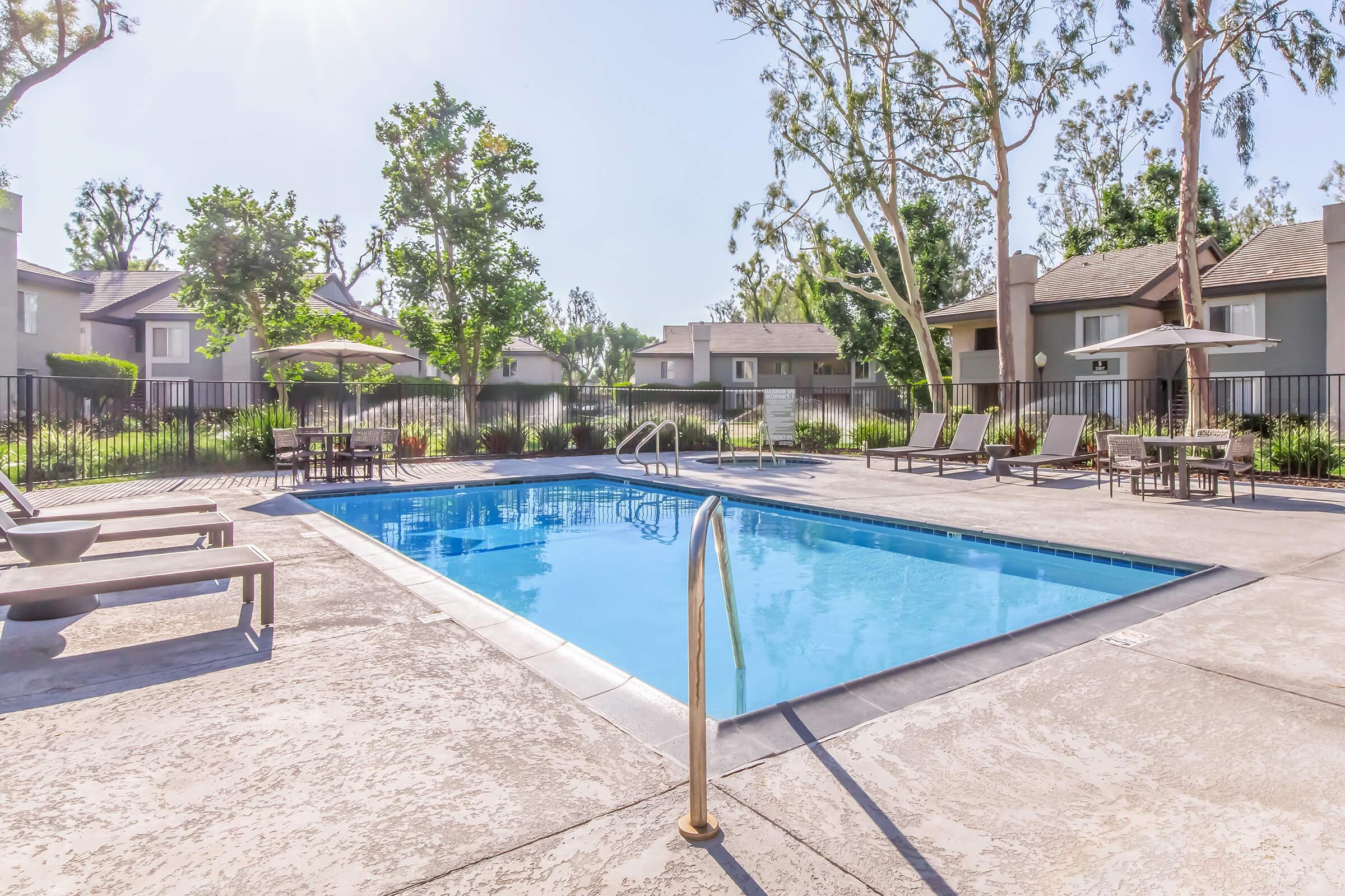 A bright outdoor swimming pool surrounded by lounge chairs and umbrellas, with trees and landscaped greenery in the background. Residential buildings are visible in the distance, creating a serene and inviting atmosphere for relaxation.