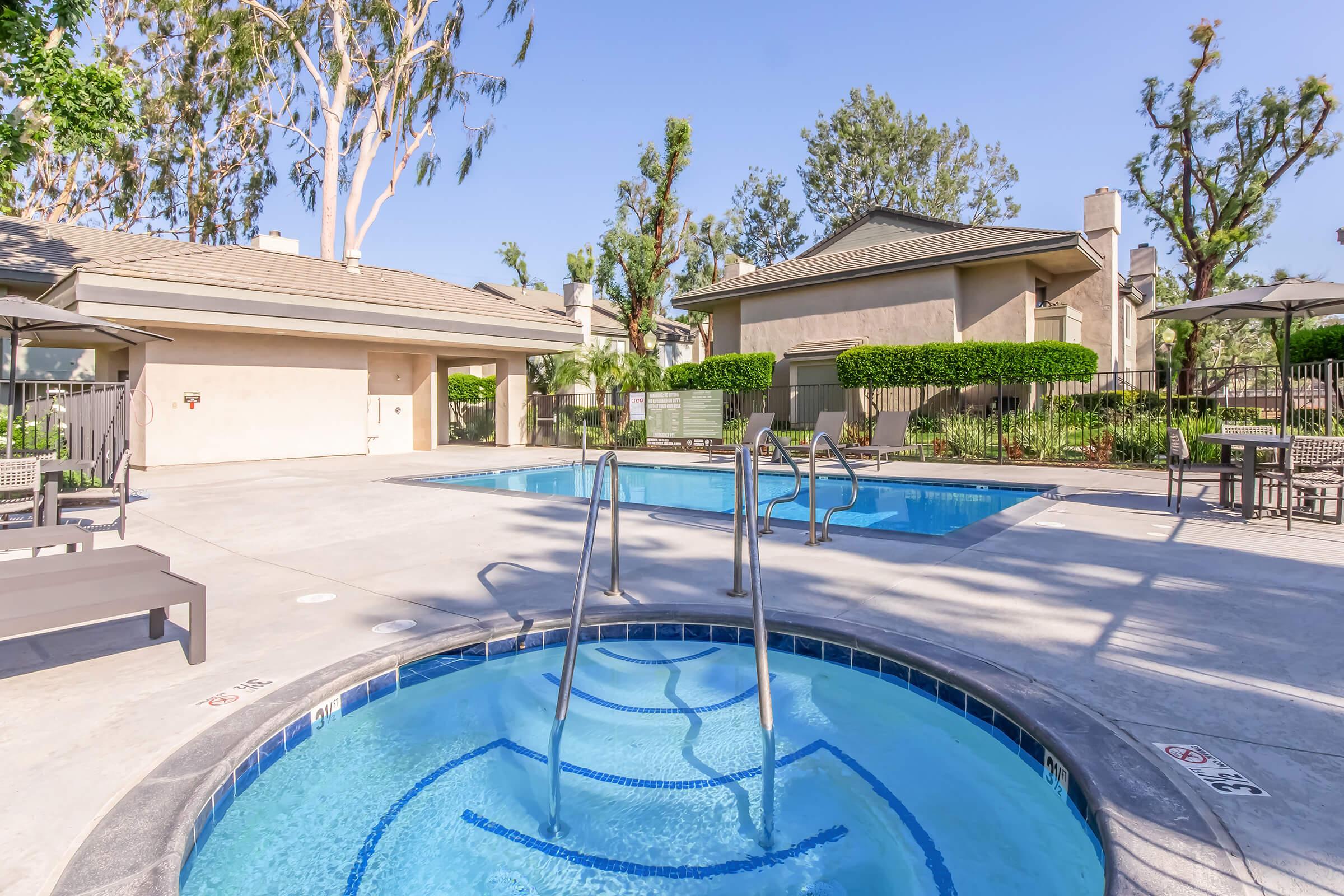 A serene outdoor pool area featuring two pools—one larger and one smaller hot tub. Surrounding the pools are lounge chairs, umbrellas, and well-maintained landscaping with trees. The sky is clear and blue, creating a peaceful setting for relaxation.