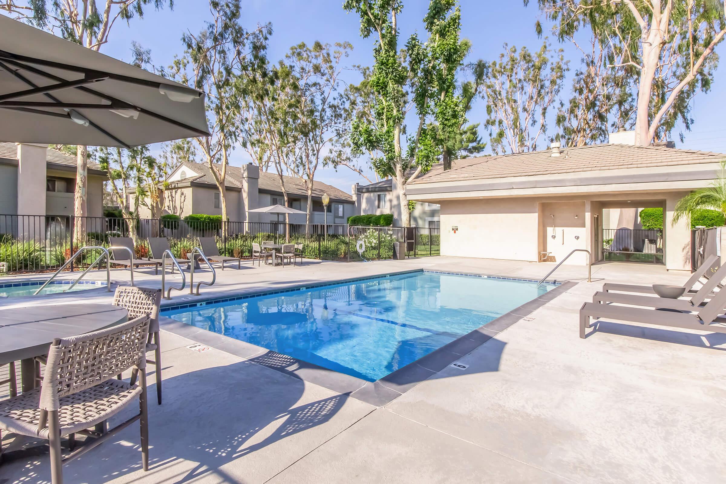 A clear blue swimming pool surrounded by lounge chairs and a table. In the background, there are trees and residential buildings with a well-maintained landscape. A covered area with an umbrella offers shade beside the pool. The setting is inviting and ideal for relaxation.