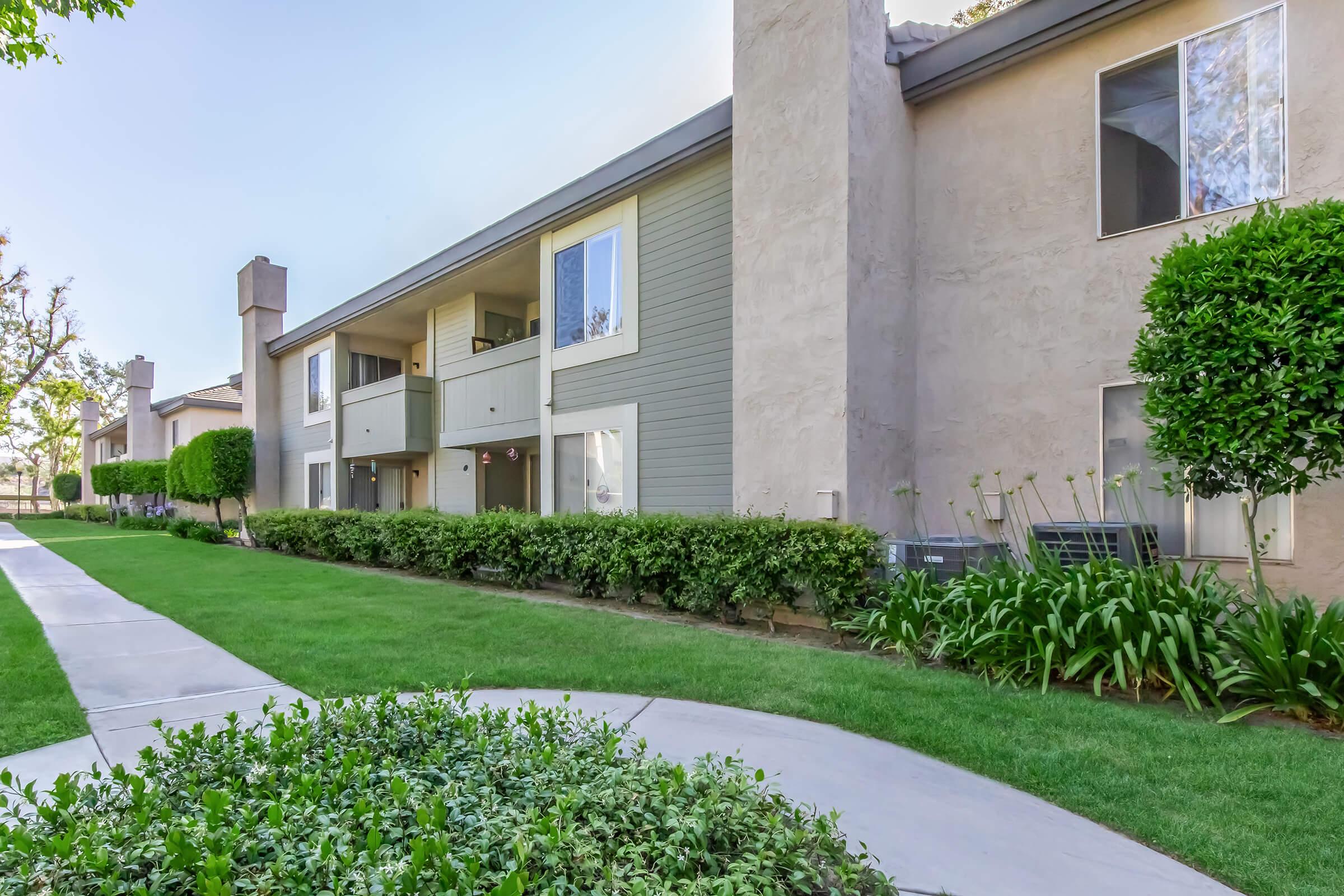 Two-story residential apartment buildings with light-colored exteriors and green landscaping. A paved walkway leads through well-maintained grass, flanked by shrubs and plants, creating a serene outdoor space. Windows on each unit reflect the surroundings, indicating a sunny day.