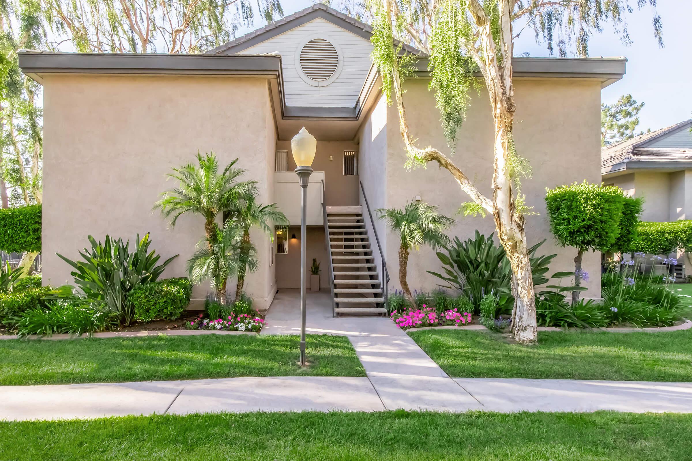 Two-story apartment building surrounded by greenery, including palm trees and flowering plants. A well-maintained pathway leads to the entrance, flanked by lawn and shrubs. A lamppost is visible in front of the building, adding to the property's inviting appearance.