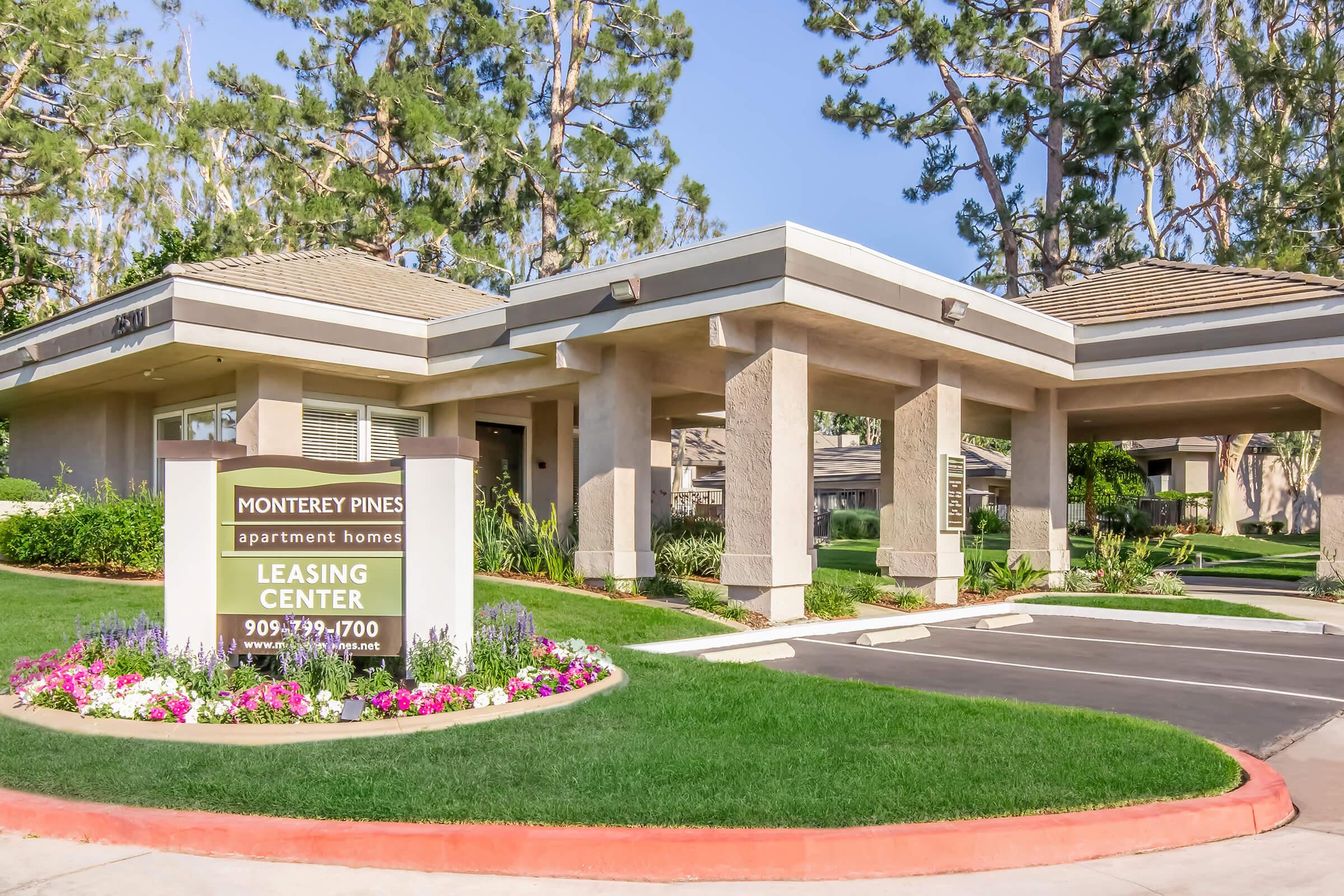 A modern leasing center for Monterrey Pines apartment homes, featuring a welcoming entrance with colorful flower beds, manicured lawns, and tall trees in the background. The building has a contemporary architectural design with a sloped roof and signage displaying contact information.