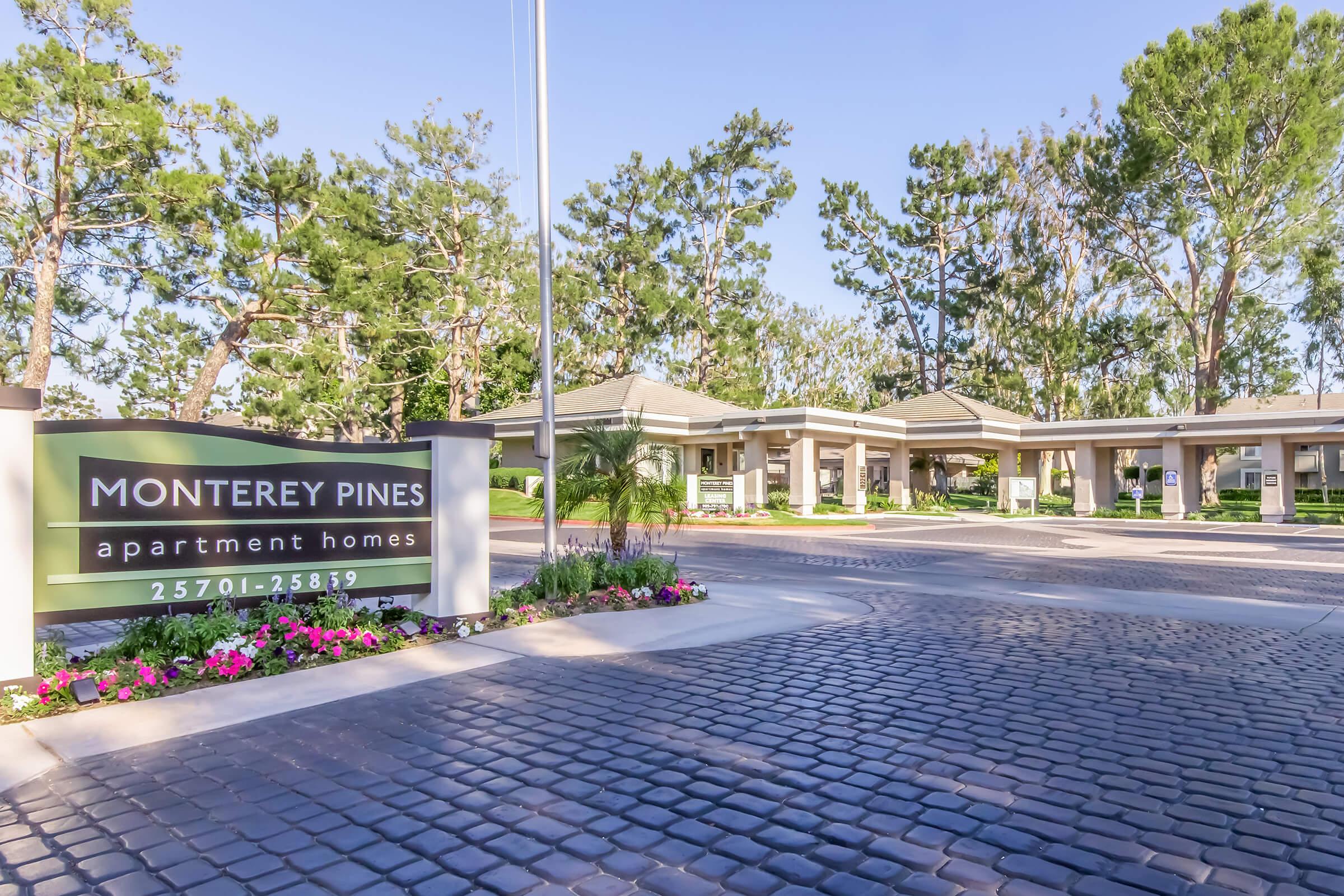 Sign for Monterey Pines apartment homes, featuring a landscaped entrance with colorful flowers and trees. The sign includes the name "Monterey Pines" prominently displayed, along with an address range. The setting is bright and inviting, suggesting a welcoming residential community.