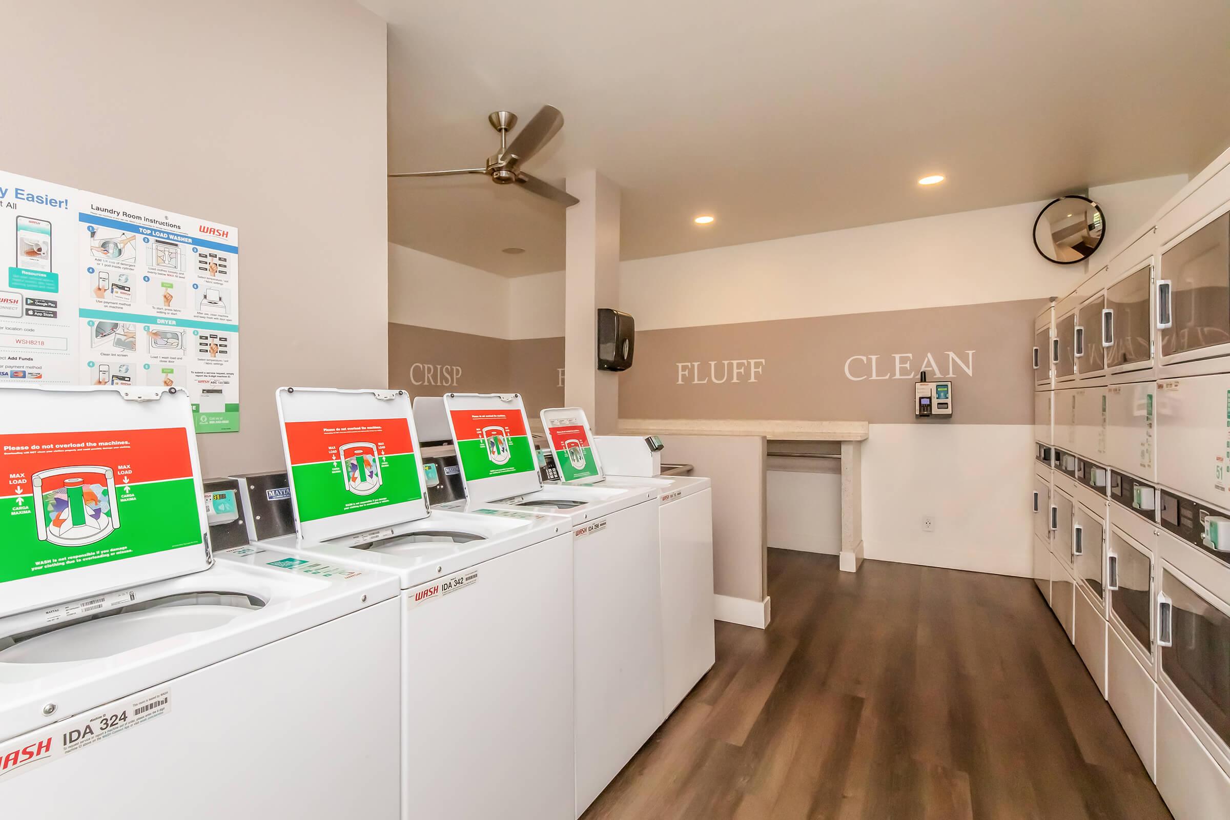 A modern laundry room featuring several washing machines with digital displays. Clean, well-lit space with a minimalist design, including laundry supplies on the wall and a folding area. The decor features the words "CRISP," "FLUFF," and "CLEAN" on the wall, enhancing the welcoming atmosphere.