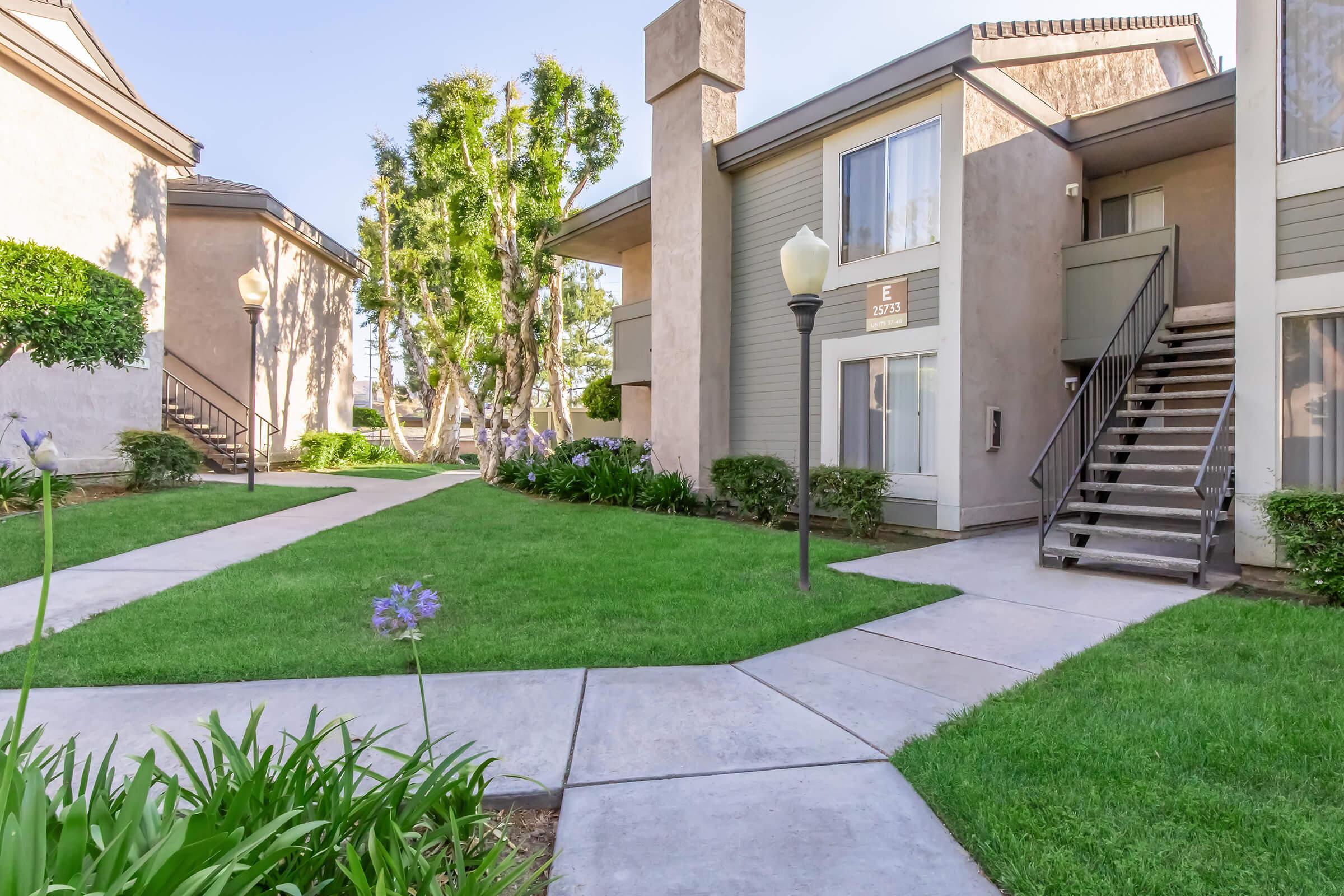 A well-maintained apartment complex featuring two-story buildings with beige exteriors, surrounded by green lawns and landscaped gardens. A paved pathway leads to the entrance of one building, with a stairway visible. Decorative street lamps are positioned along the walkway, enhancing the inviting atmosphere.
