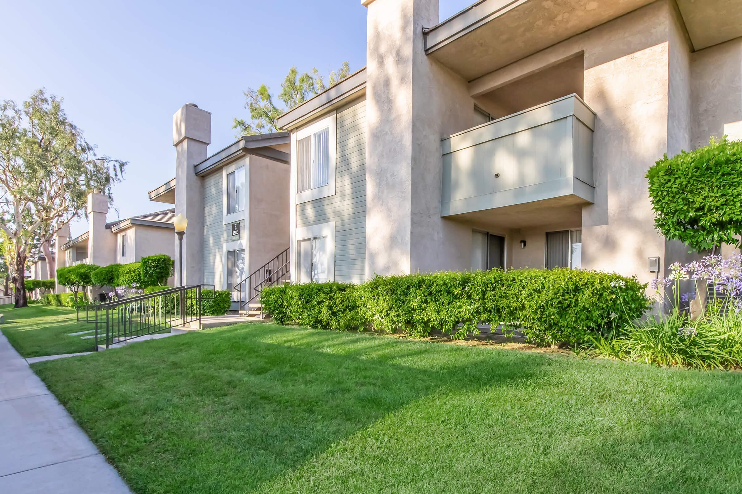 Exterior view of a multi-unit residential building featuring beige and gray siding, with balconies and landscaped green lawns. The pathway leads to the entrance, surrounded by well-maintained shrubs and grass, under a clear blue sky.