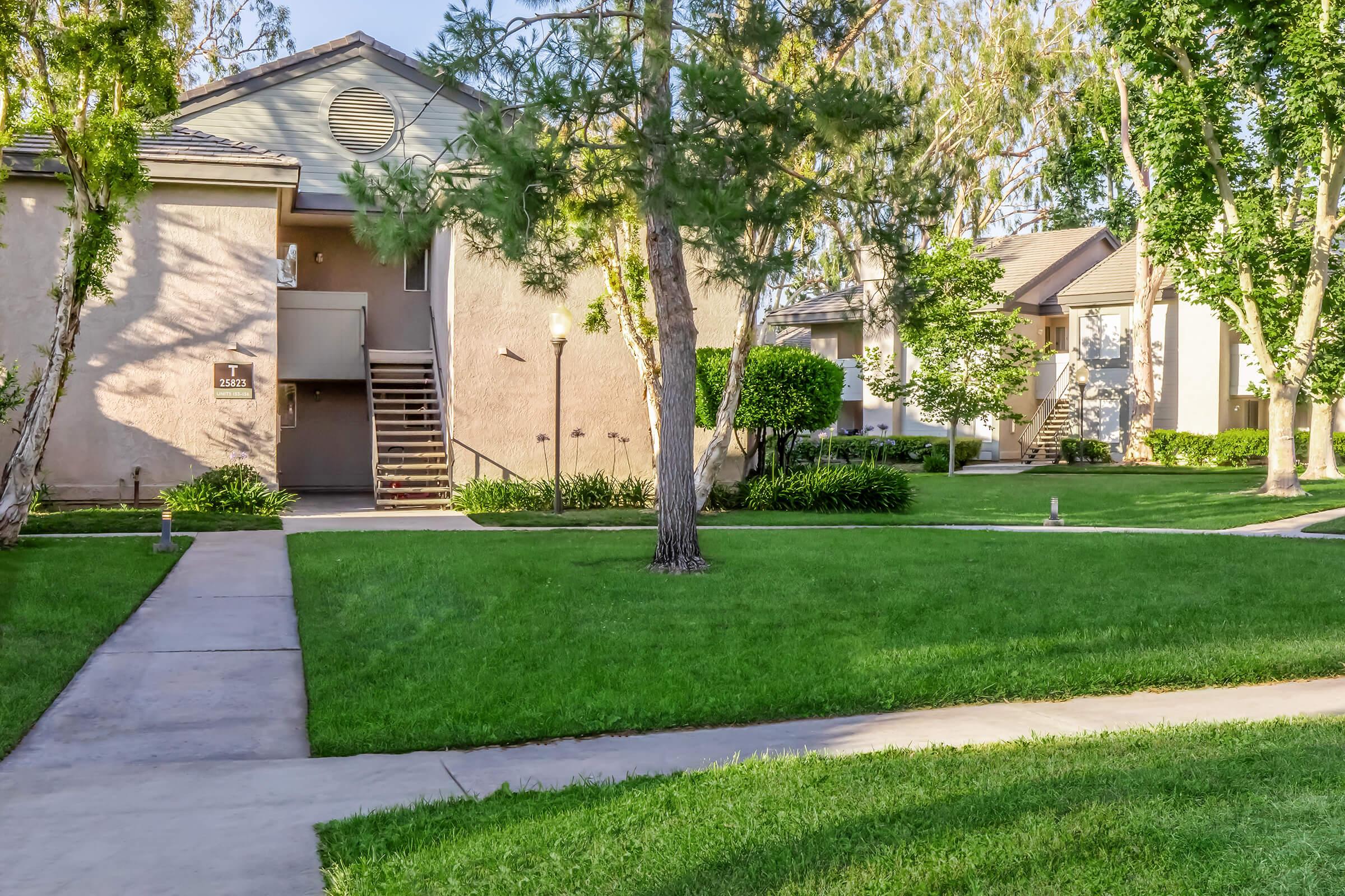 A well-maintained residential area featuring two apartment buildings, lush green lawns, and neatly trimmed shrubs. A paved walkway leads up to the buildings, with trees providing shade, creating a pleasant outdoor environment.