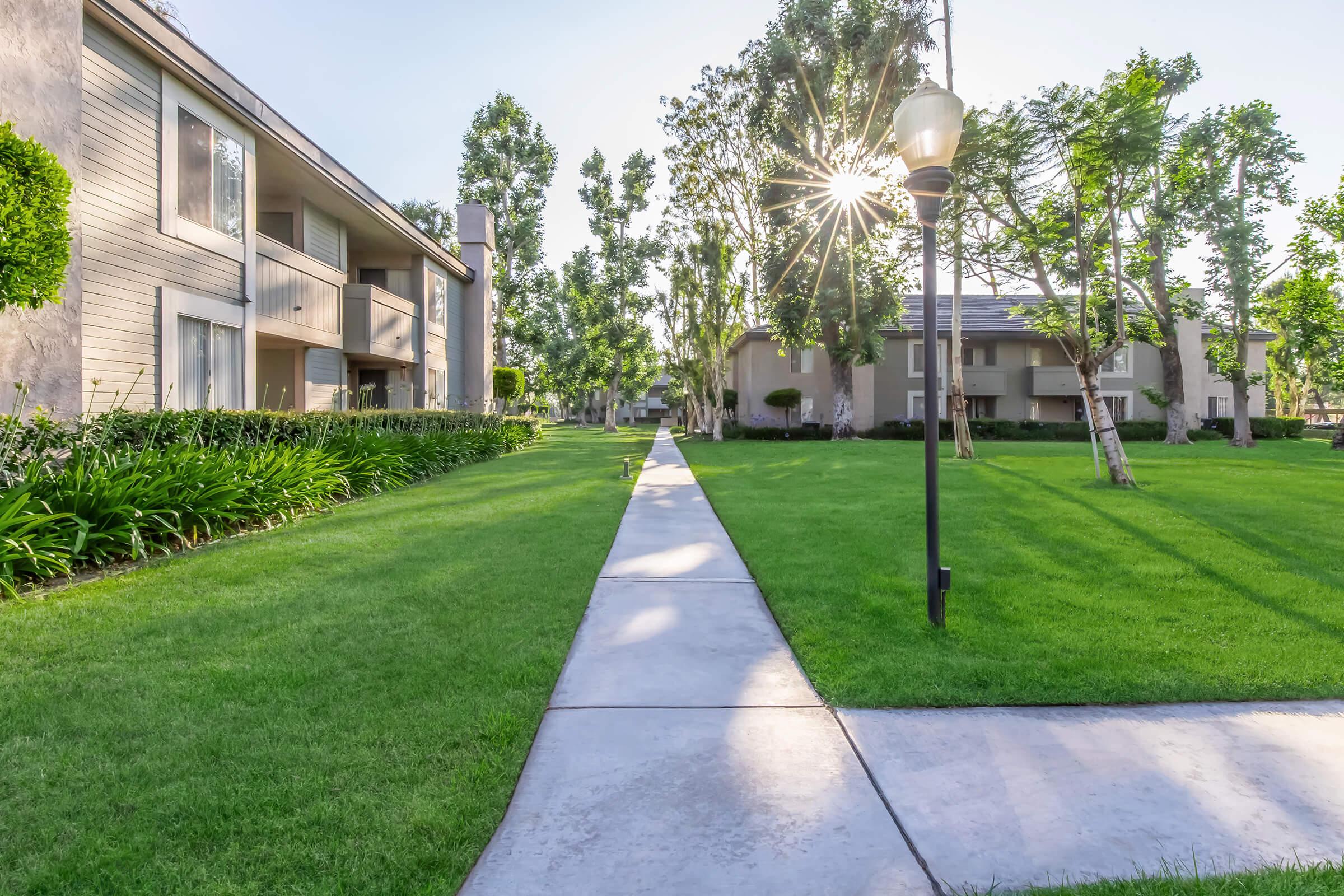 A well-maintained pathway lined with lush green grass leads through a residential area featuring two apartment buildings. Sunlight filters through the trees, creating a warm, inviting atmosphere. A lamppost stands along the path, enhancing the aesthetic of the community.