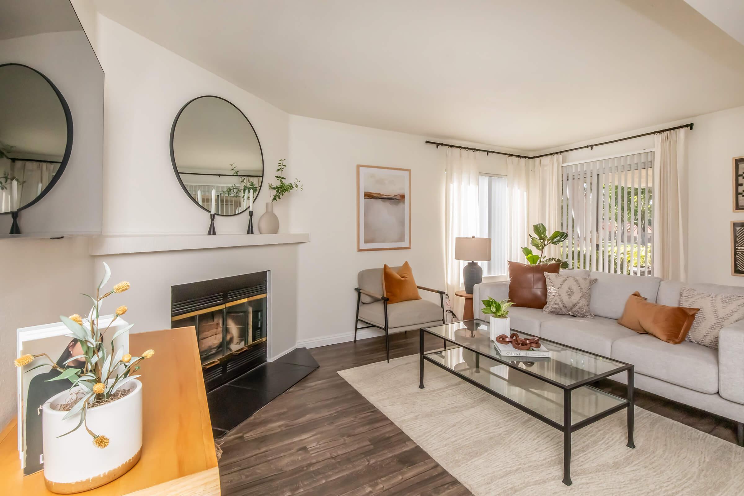 A cozy living room featuring a light gray sofa with orange and brown cushions, a glass coffee table, and a stylish fireplace. There's a round mirror above the mantel, soft curtains, and decorative plants adding a touch of greenery. The space is bright and welcoming, with modern decor elements.