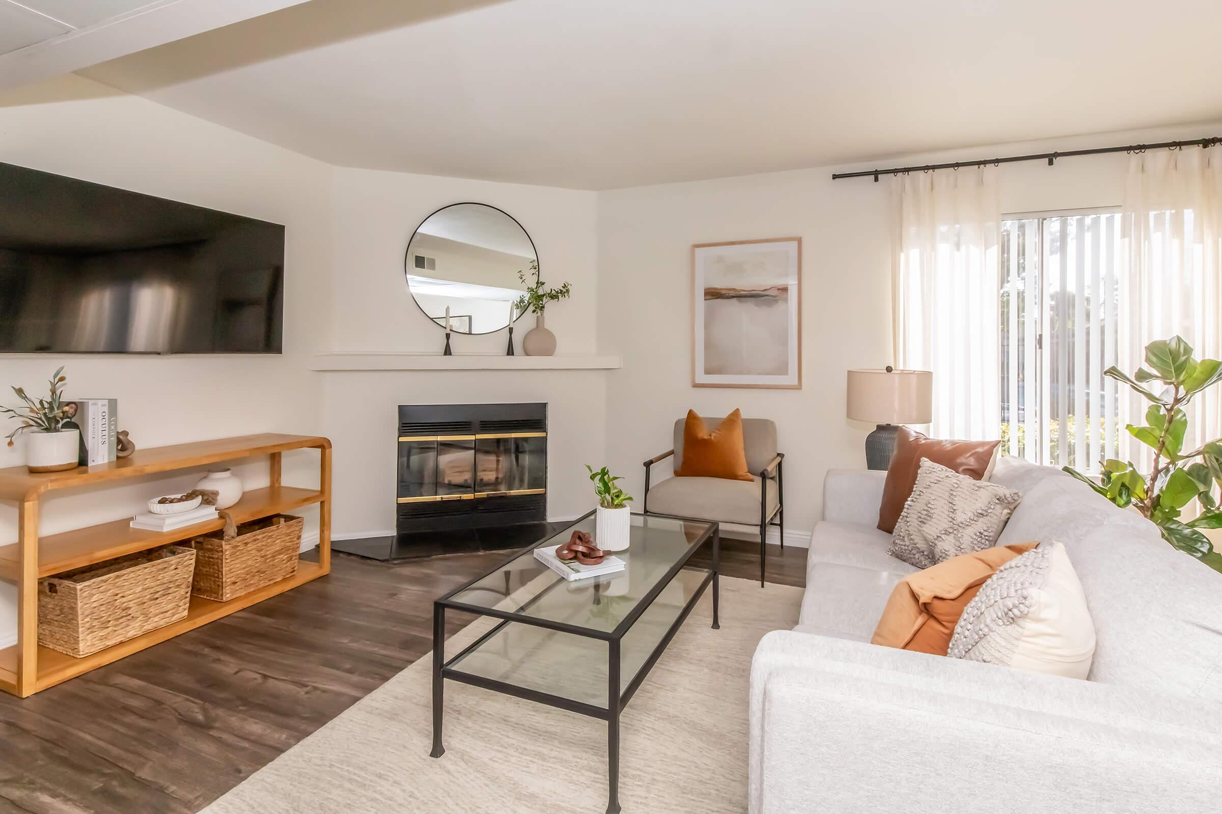 A cozy living room featuring a light gray sofa with decorative pillows, a glass coffee table, and a wooden shelf with storage baskets. A modern TV hangs above a fireplace, and a large round mirror complements the space. Natural light streams in through sheer curtains, enhancing the warm and inviting atmosphere.