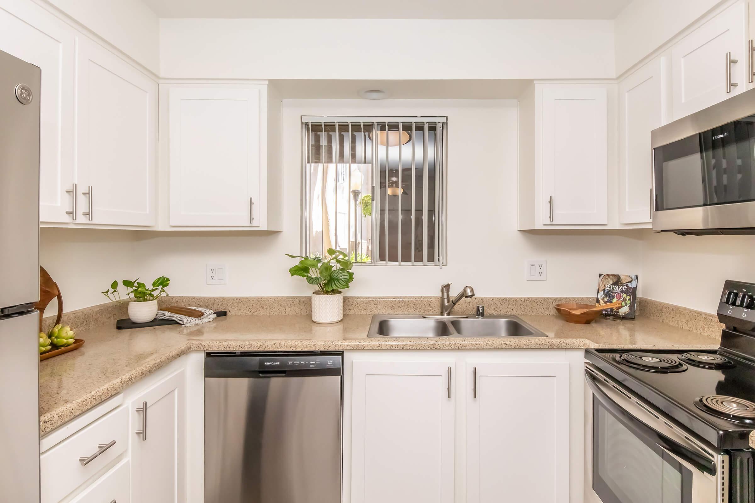 A modern kitchen featuring white cabinets, a stainless steel dishwasher, and a sink with a window above it. The countertop is beige with a few decorative plants and kitchen items. An oven and microwave are also visible on the right side. Bright lighting enhances the clean, airy feel of the space.