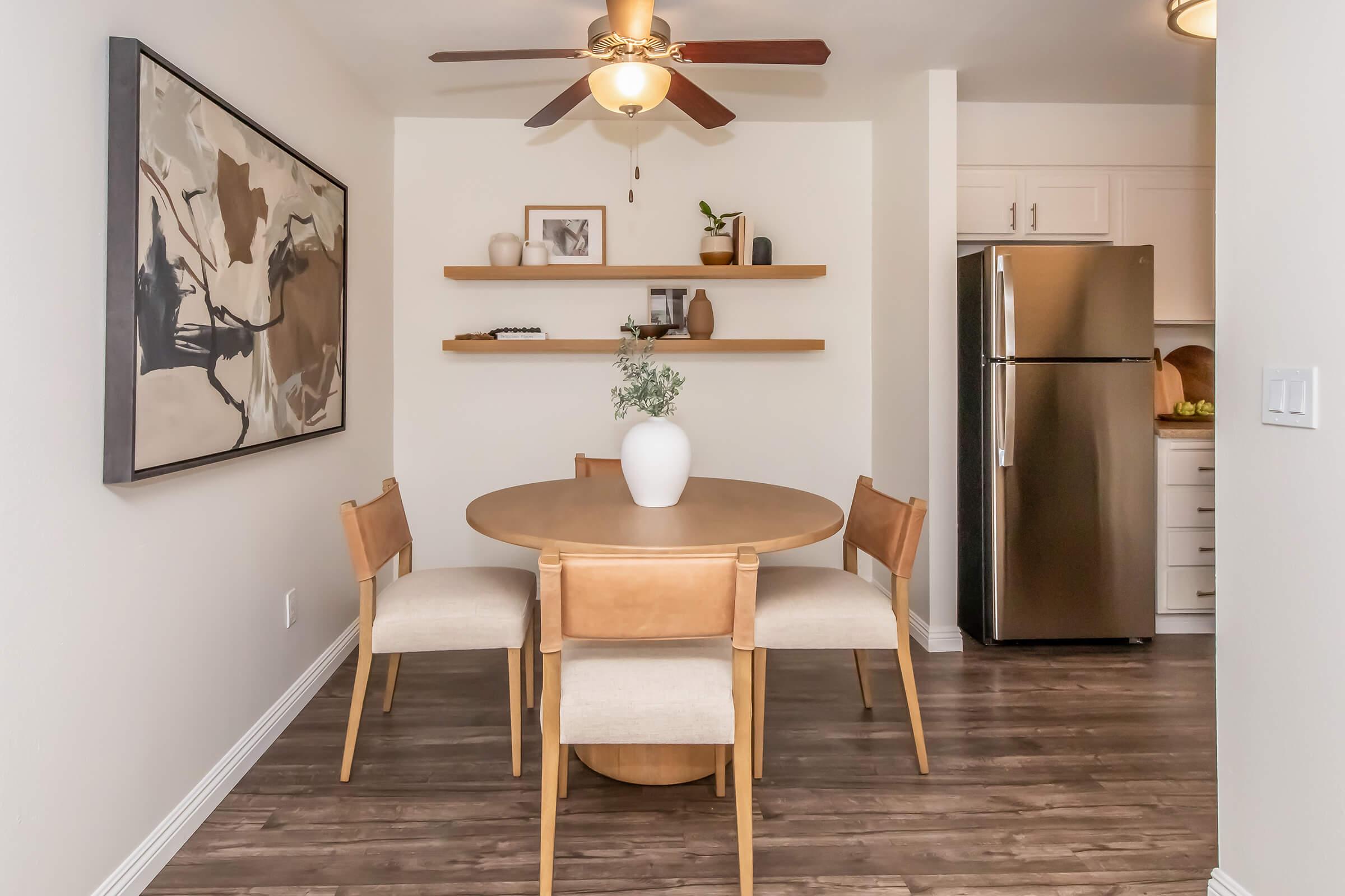 A modern dining area featuring a round wooden table surrounded by four light-colored chairs. A large abstract painting hangs on the wall, and a shelf showcases decorative items. A ceiling fan is visible, along with a stainless steel refrigerator in the background, contributing to a stylish, inviting atmosphere.