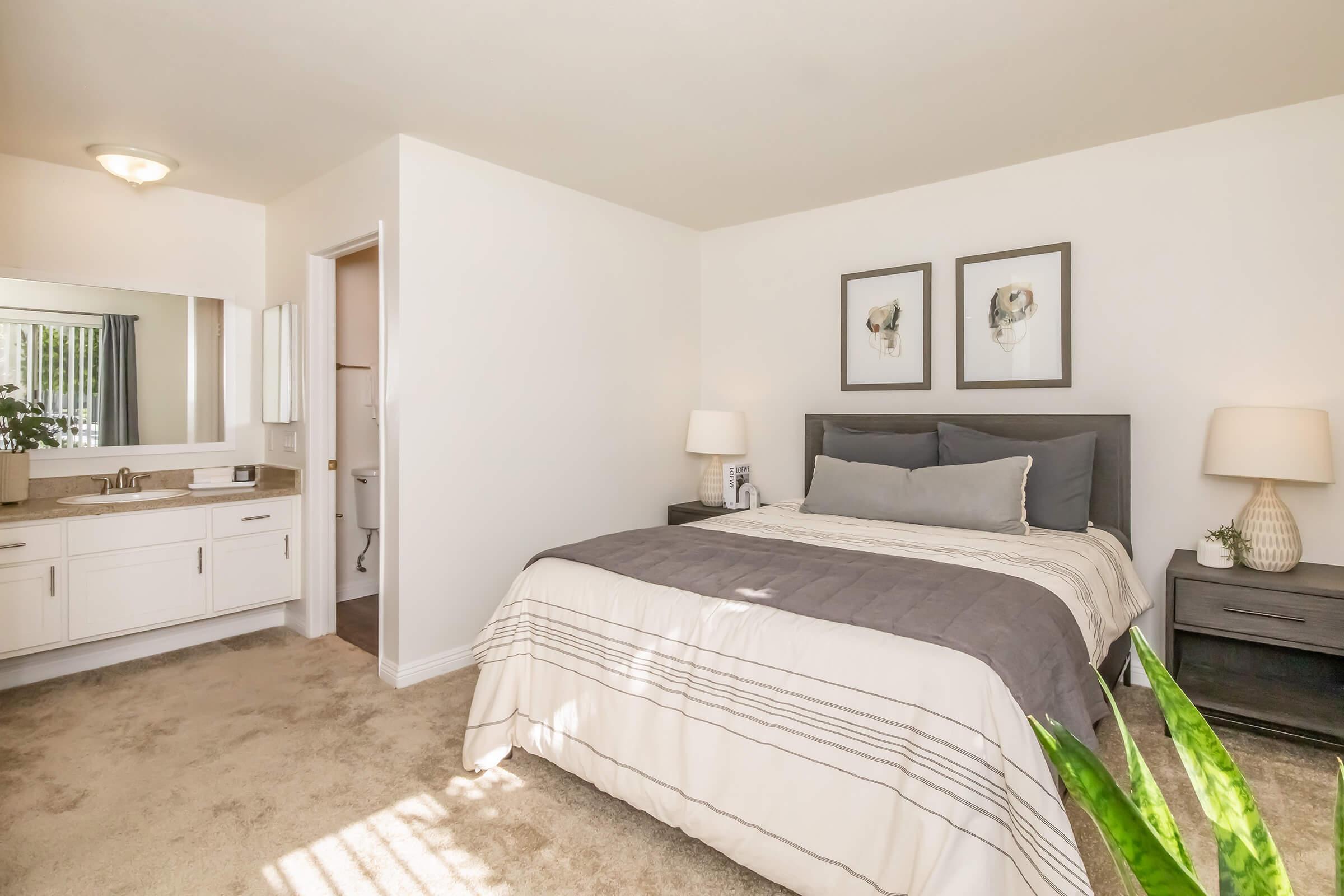 Cozy bedroom featuring a queen-sized bed with gray bedding, two nightstands with lamps, wall art of floral prints, a dresser, and a view into a bathroom. Soft light filters through the window, illuminating the neutral color palette and carpeted flooring. Green plant adds a touch of nature to the space.