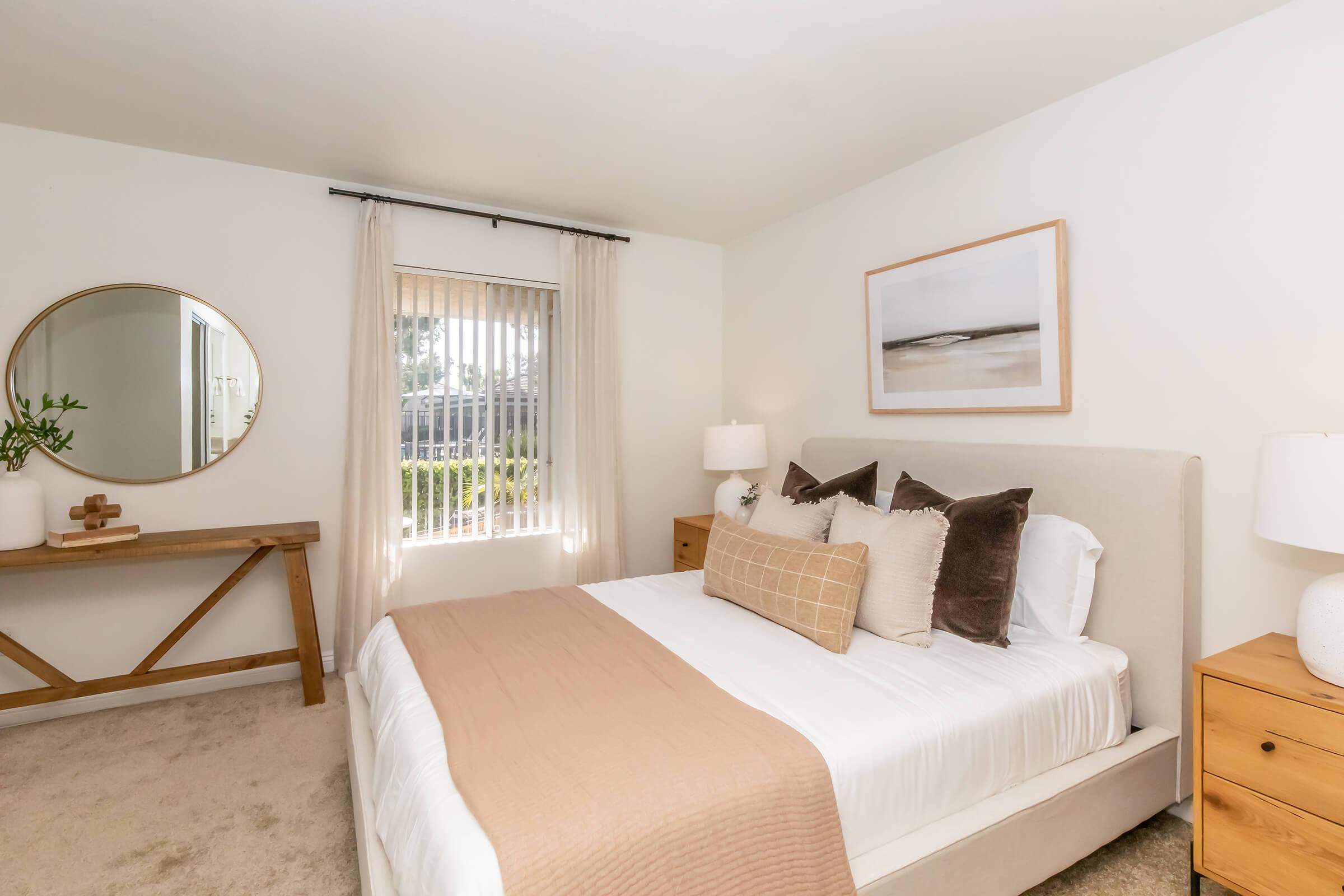 A cozy bedroom featuring a neutral color palette. A comfortable bed with decorative pillows is centered in the space. There is a wooden nightstand on each side, and a round mirror above a console table. Natural light streams through a window with sheer curtains, creating a warm and inviting atmosphere.