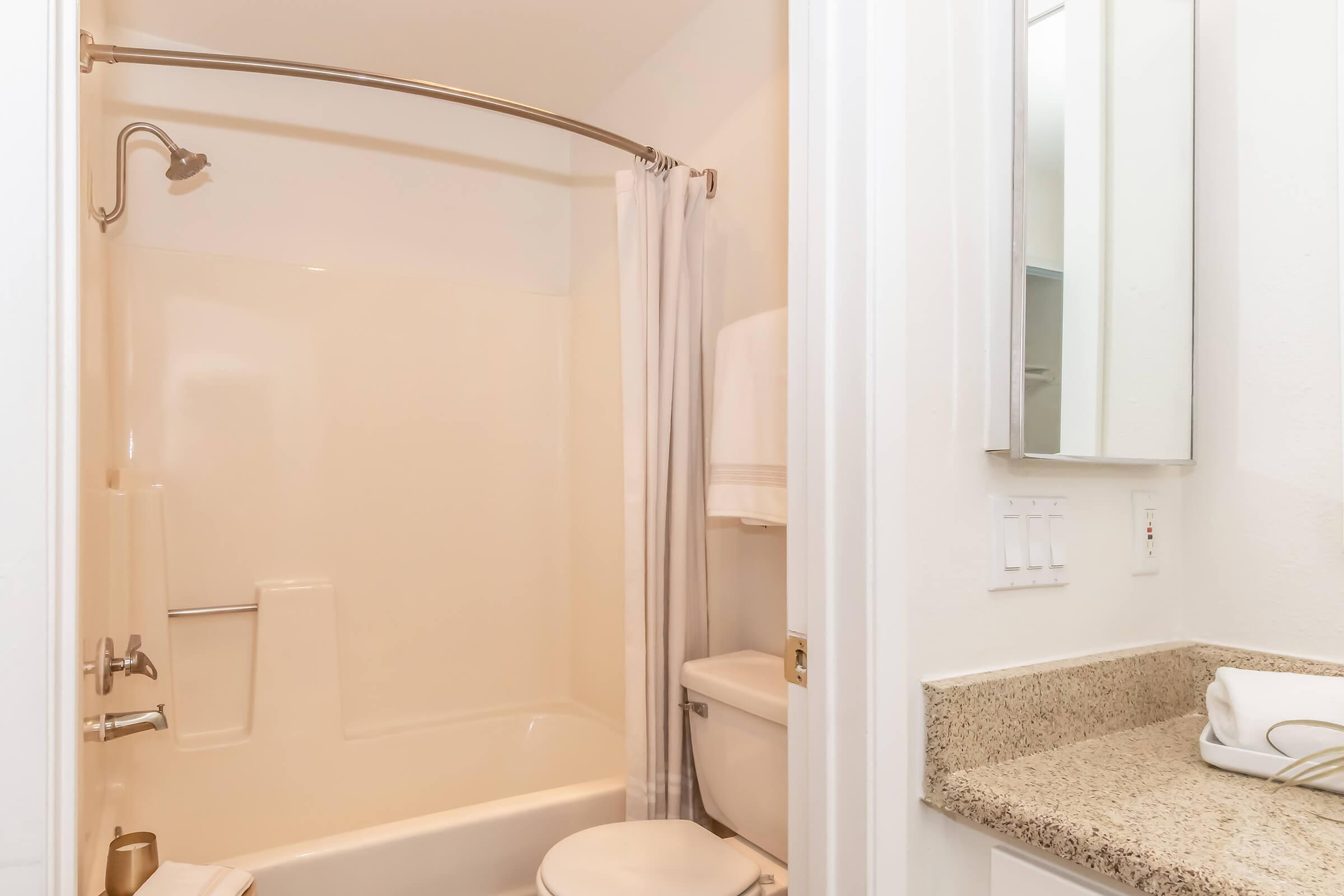 A clean, modern bathroom featuring a shower with a curved curtain rod, a bathtub, a toilet, and a sink with a granite countertop. The walls are light-colored, and there are white towels hanging nearby. A mirror is mounted above the sink.