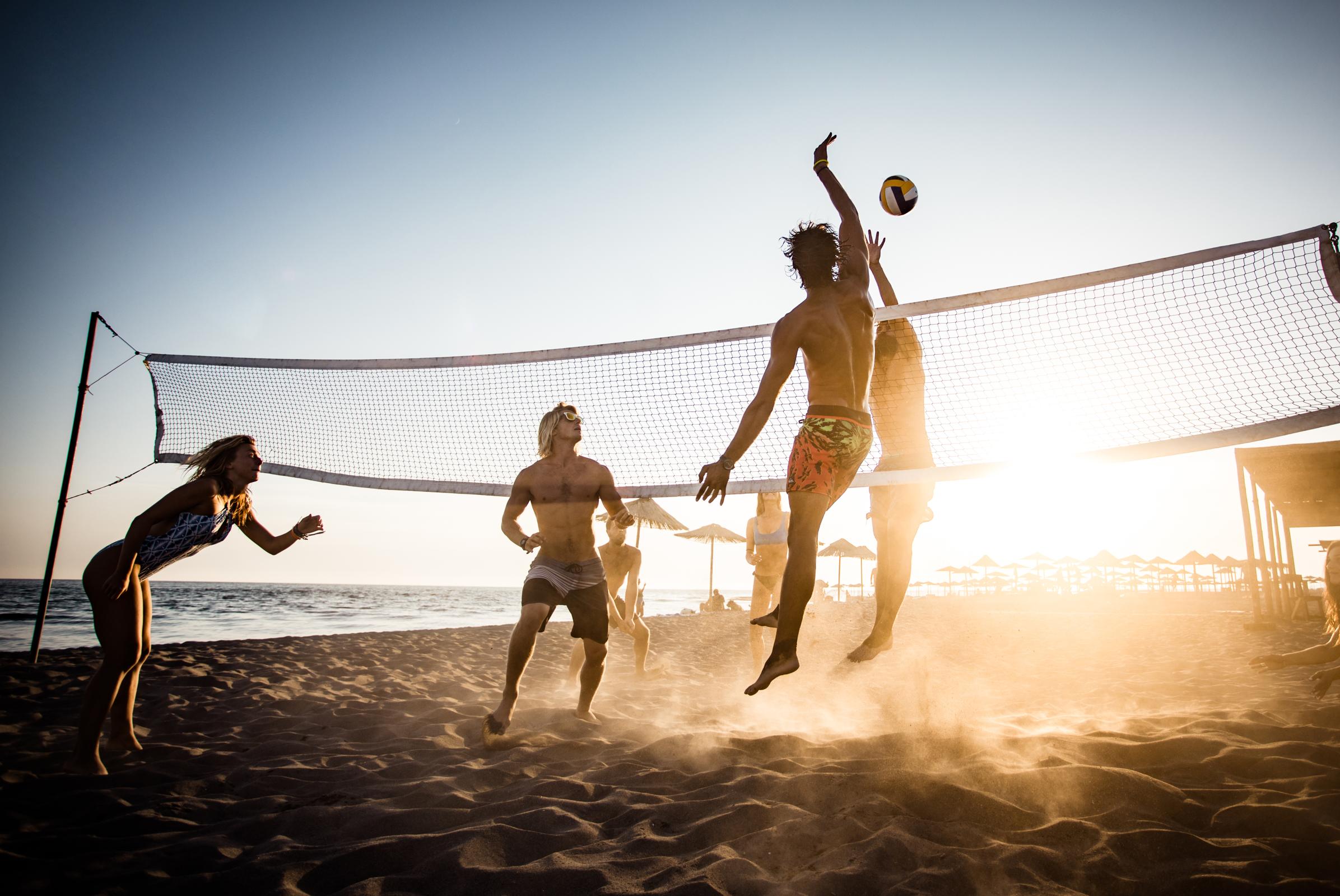A group of four people playing beach volleyball at sunset, with two players in the air attempting to hit the ball. The scene captures a sandy beach with a net in the foreground and the sun setting over the water in the background, creating a warm, vibrant atmosphere.