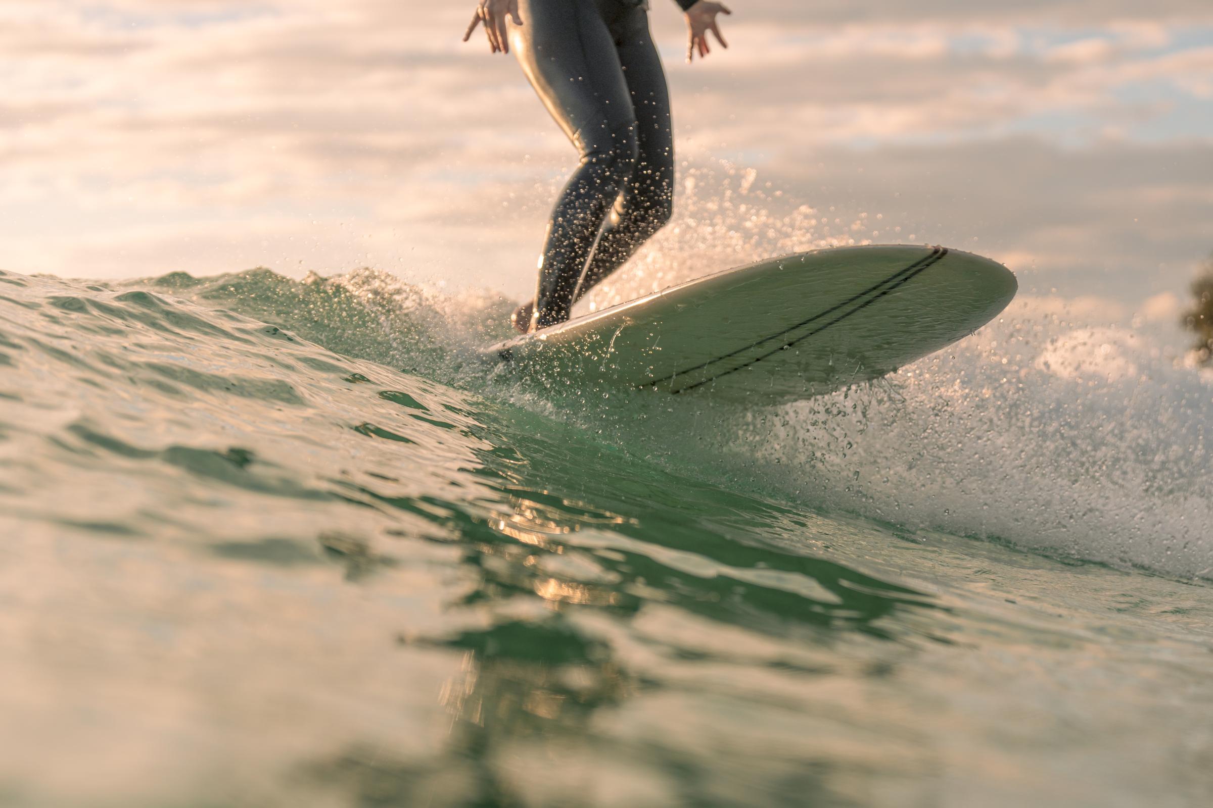 A surfer riding a wave during golden hour, with a focus on their lower body and surfboard, which is cutting through the water. Soft waves and a shimmering sea create a dynamic and serene atmosphere. The background features a cloudy sky, enhancing the tranquil yet exhilarating scene.