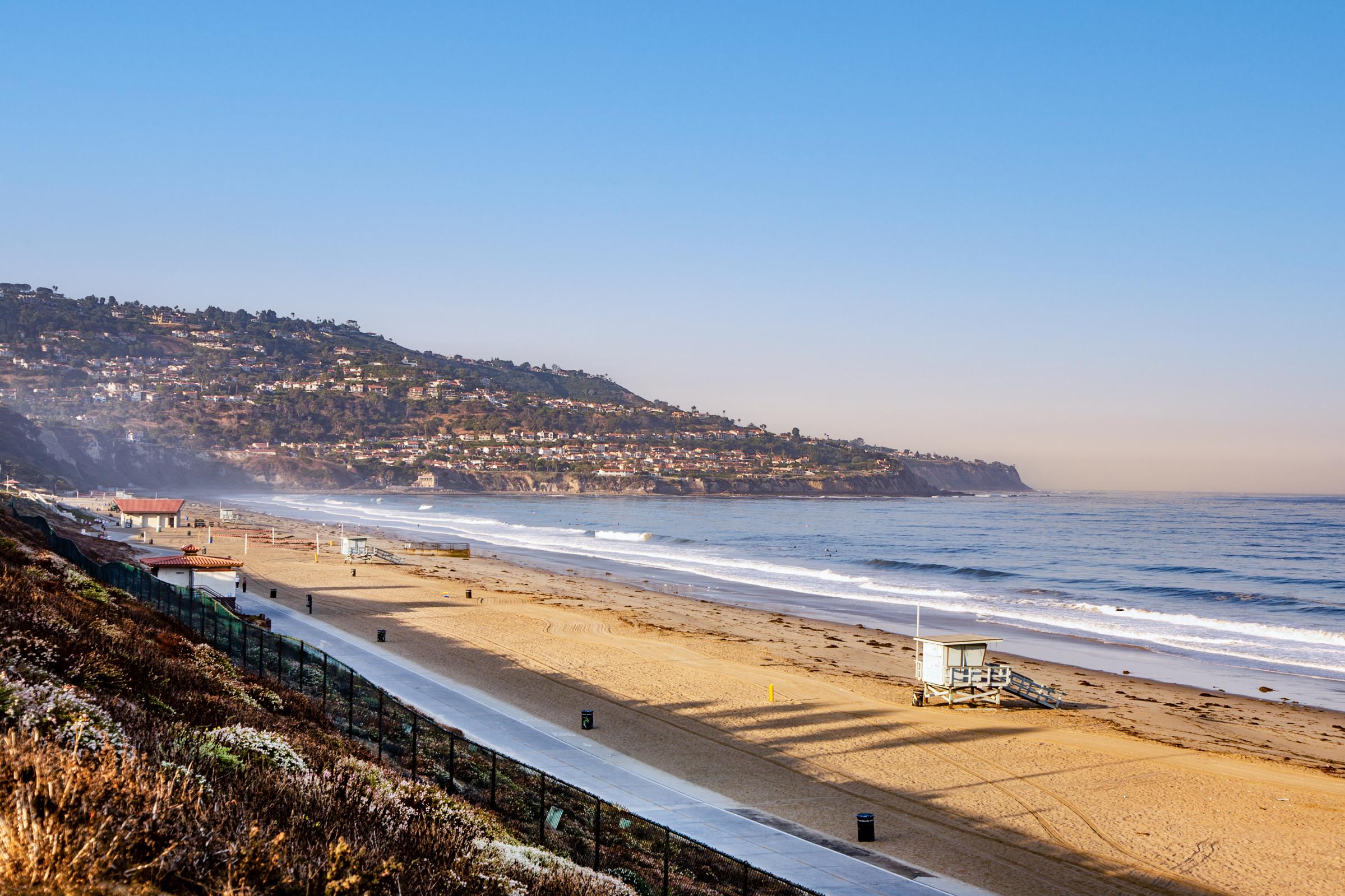 A serene beach scene with gentle waves lapping at the shore. The sandy beach is lined with a walking path and a lifeguard tower in the foreground. In the background, a coastal hillside is dotted with homes, under a clear blue sky. The atmosphere conveys a peaceful morning at the beach.