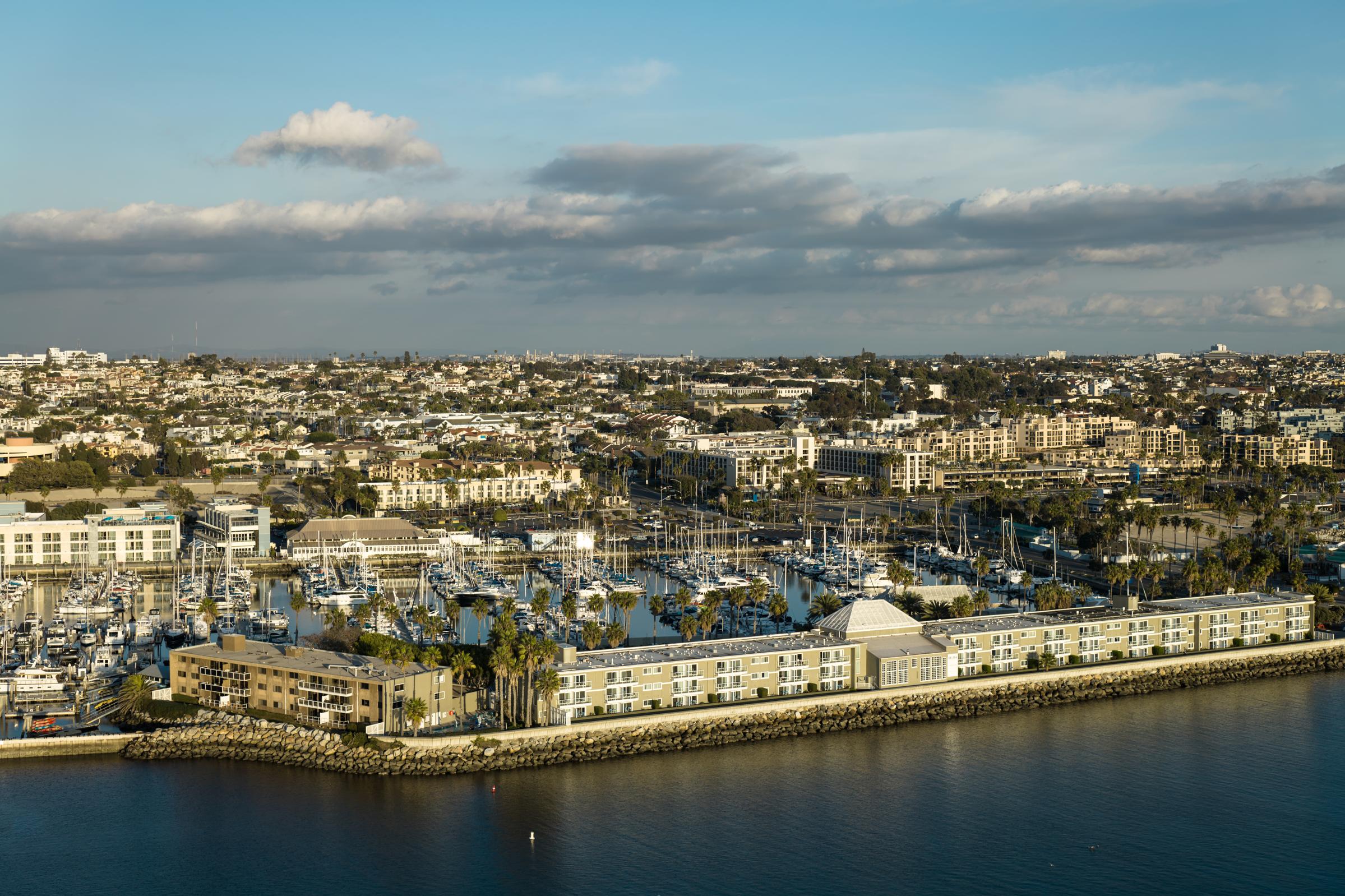 Aerial view of a coastal marina with numerous boats docked. In the foreground, a modern waterfront building is visible along the shoreline, while behind it, a sprawling urban landscape with residential and commercial structures extends into the distance under a blue sky with scattered clouds.