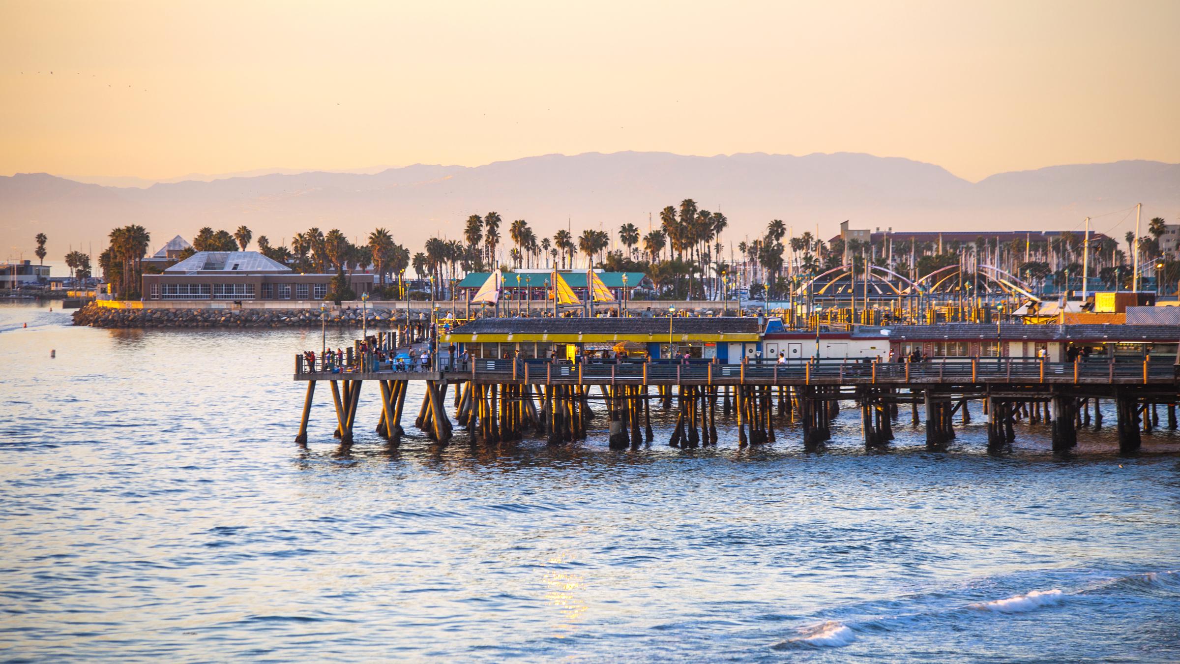 A vibrant pier extends over calm waters during sunset, with palm trees lining the shore. The scene includes attractions and people enjoying the seaside atmosphere, with soft, warm light illuminating the landscape and mountains in the background. The overall mood is relaxed and picturesque.