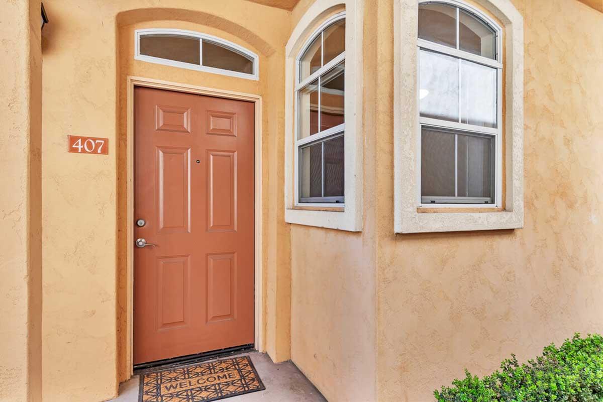 A close-up view of a decorative entryway featuring an orange-brown door marked with the number 407. A small welcome mat is positioned at the base of the door, and light-colored stucco walls frame the entrance. Windows beside the door allow natural light to enter the space.