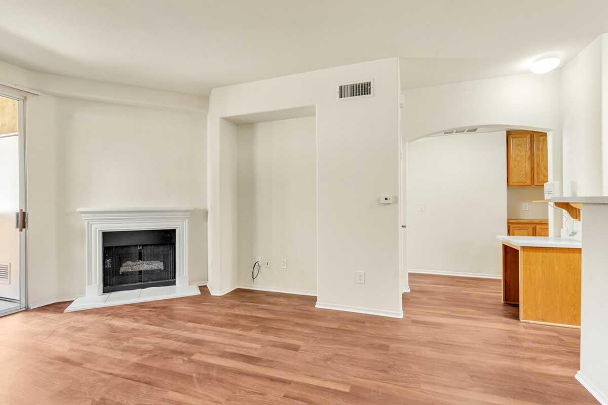 A spacious, empty living room featuring light-colored walls and hardwood flooring. On one side, there is a white fireplace and an archway leading to a kitchen area with wooden cabinets. Large windows allow natural light to fill the space, creating a bright and airy atmosphere.