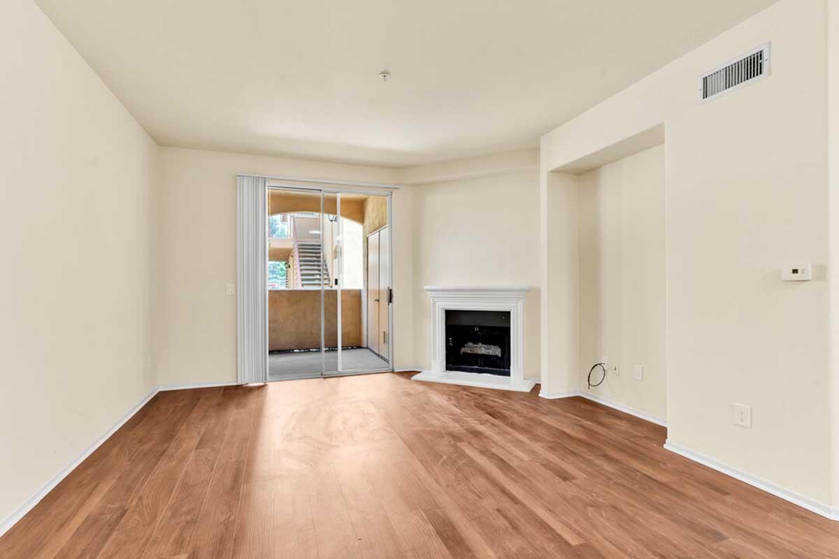 A spacious, empty living room featuring light-colored walls, wooden flooring, and a fireplace. Large sliding glass doors provide access to an outdoor area, while a staircase is visible outside. The room has a clean and modern aesthetic, with ample natural light flooding in.
