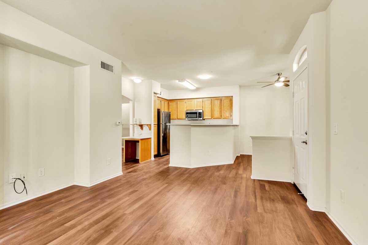 Spacious modern living area with light hardwood flooring, featuring an open kitchen with wooden cabinets, stainless steel appliances, and a breakfast bar. There is a ceiling fan, white walls, and a front door leading outside. Natural light brightens the room, creating a welcoming atmosphere.