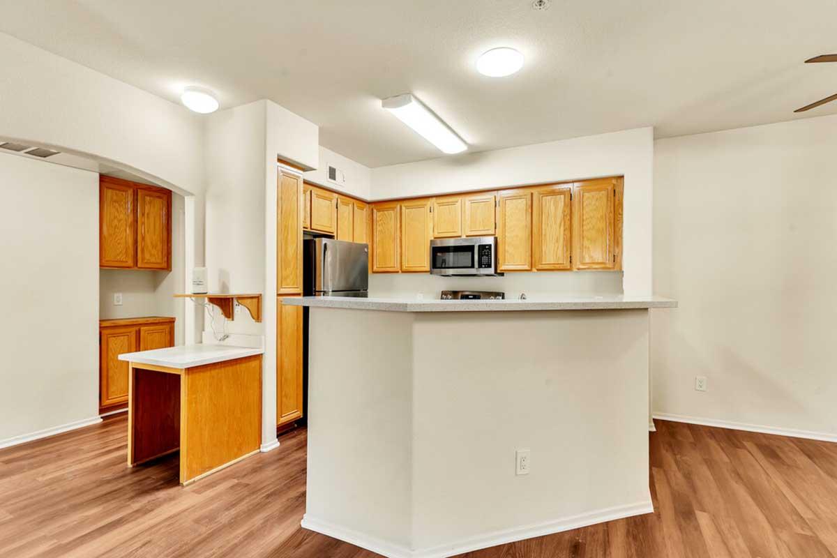 A modern kitchen featuring light wooden cabinetry, stainless steel appliances, and a countertop with bar seating. The space has a warm, inviting atmosphere with neutral-colored walls and wood flooring, illuminated by overhead lighting. A small countertop extension is visible, creating additional workspace.
