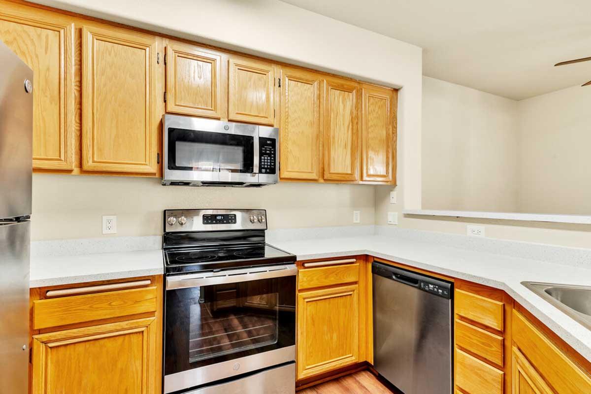 Modern kitchen featuring wooden cabinets, a stainless steel refrigerator, a microwave above a sleek black oven, and a dishwasher. The countertops are light-colored, and the space is well-lit with neutral walls and a ceiling fan visible in the background.