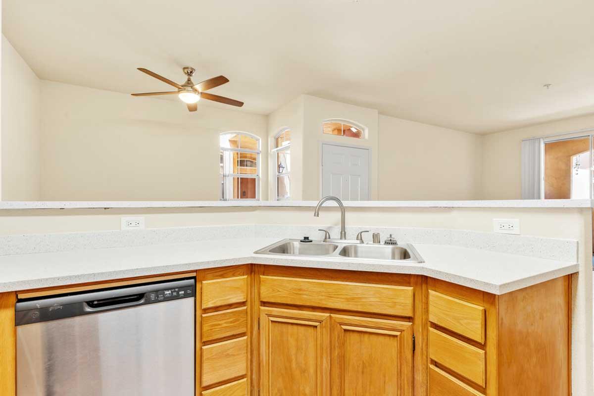 A bright kitchen featuring light-colored walls, a ceiling fan, and wood cabinetry. The countertop has a dual sink and a dishwasher. Large windows allow natural light, complemented by an open layout leading to another room.