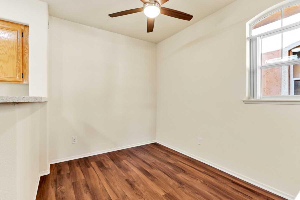 A small, empty room featuring light-colored walls, a wooden floor, and a ceiling fan. There is a window with white trim allowing natural light in, and a section of a countertop along one wall. The space appears clean and ready for furniture or decoration.