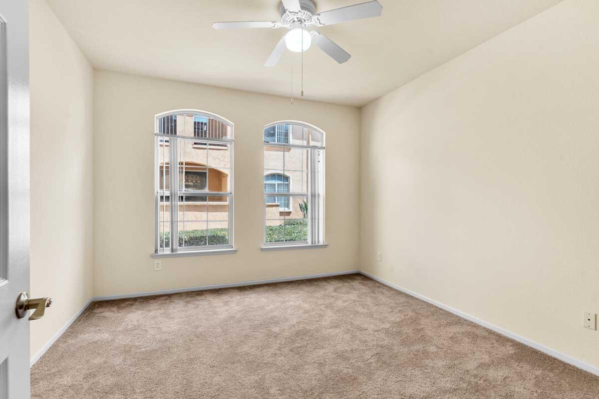 A vacant, softly lit room with beige walls and plush carpet. It features two arched windows with white trim, letting in natural light. A ceiling fan is mounted above, and the room is unfurnished, creating a spacious and inviting atmosphere.