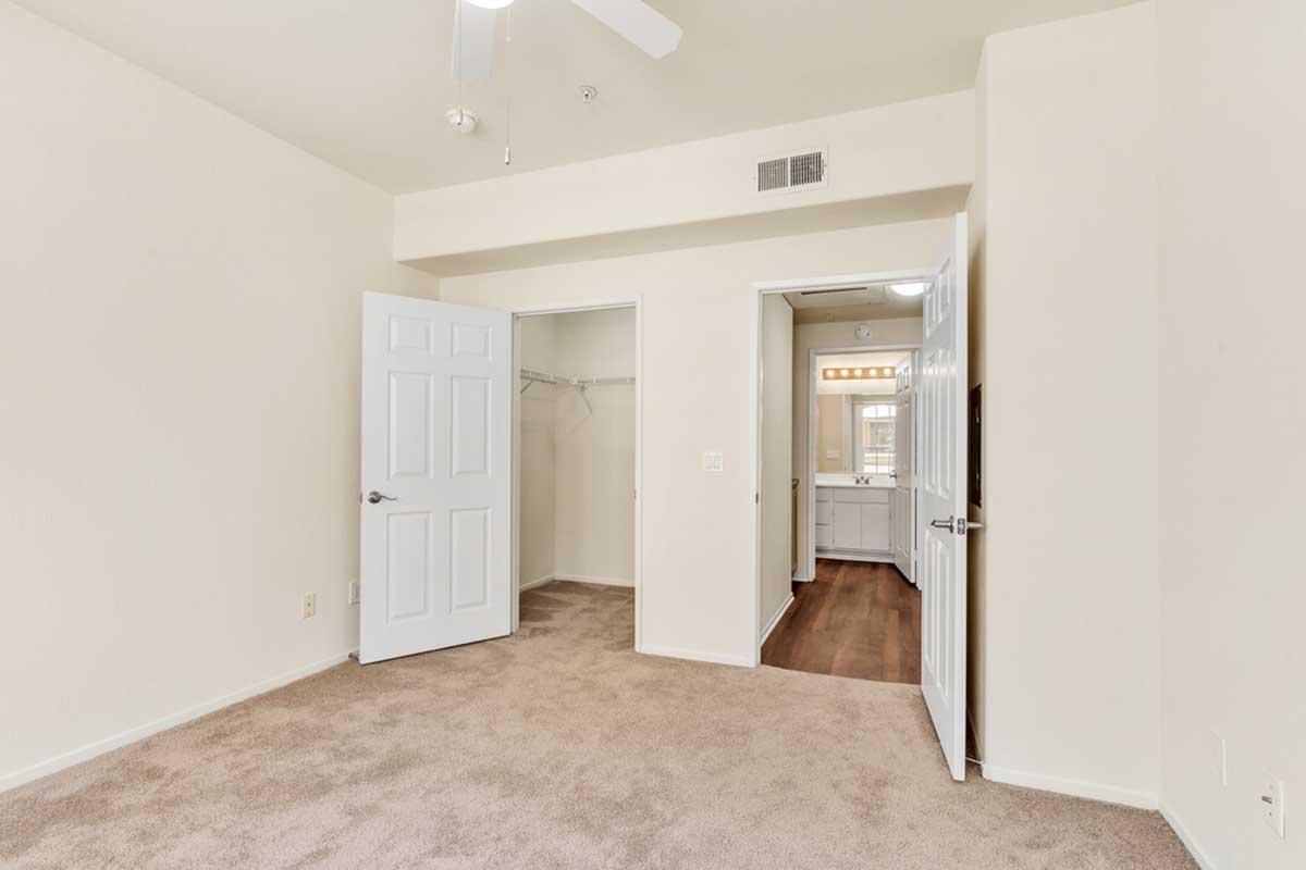 A spacious, empty bedroom featuring light beige walls and carpet. There are two open doors: one leading to a closet with no visible contents, and another to a bathroom with vanity lights visible. A ceiling fan hangs above, and natural light fills the room, creating a calm and inviting atmosphere.