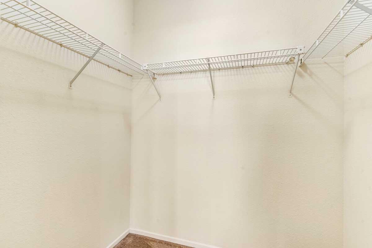 Empty closet with white wire shelving on the back wall and three hanging rods. The walls are painted light beige, and the floor is carpeted in a neutral color. The space is well-lit and appears clean and organized, suitable for storage.