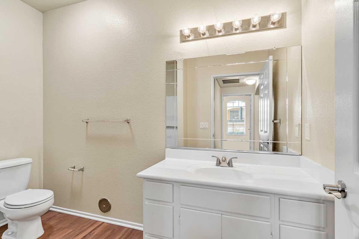 A well-lit bathroom featuring a white vanity with a sink, a large mirror above it, and a toilet. The walls are painted in a light color, and there is a light fixture above the mirror. A doorway in the background leads to another room, showing a glimpse of the interior layout.