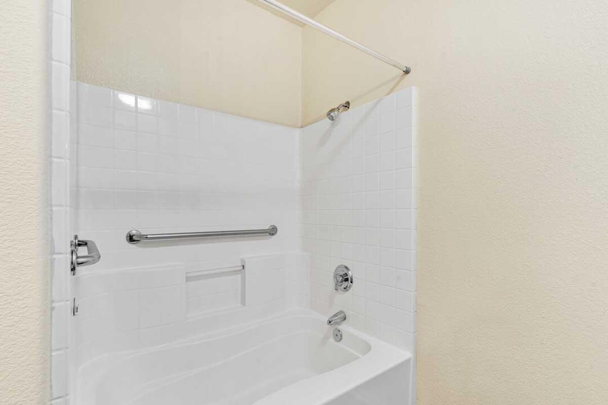 A clean, white bathroom featuring a bathtub with a showerhead above. The walls are tiled, and there is a grab bar installed for safety. The space is well-lit with a light-colored wall, creating a simple and functional design.