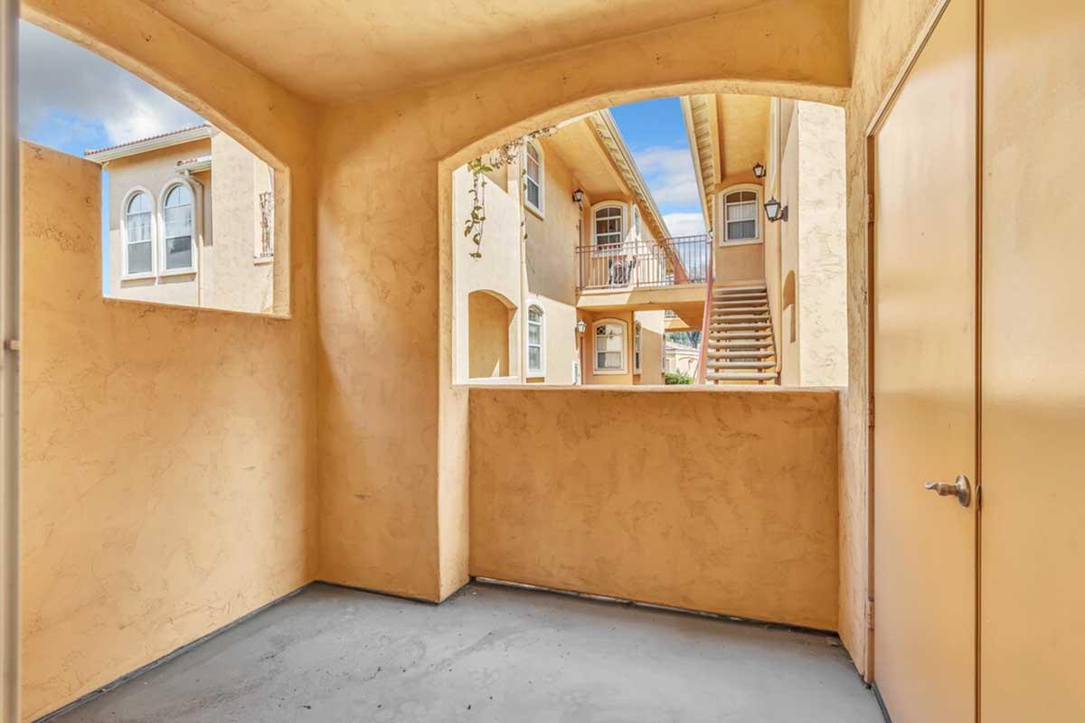 An interior view of a balcony or porch area featuring a textured, warm-colored wall. There are archways that lead to an open courtyard with stairs and additional doors visible. Natural light filters in from the outside, creating a bright, inviting atmosphere.