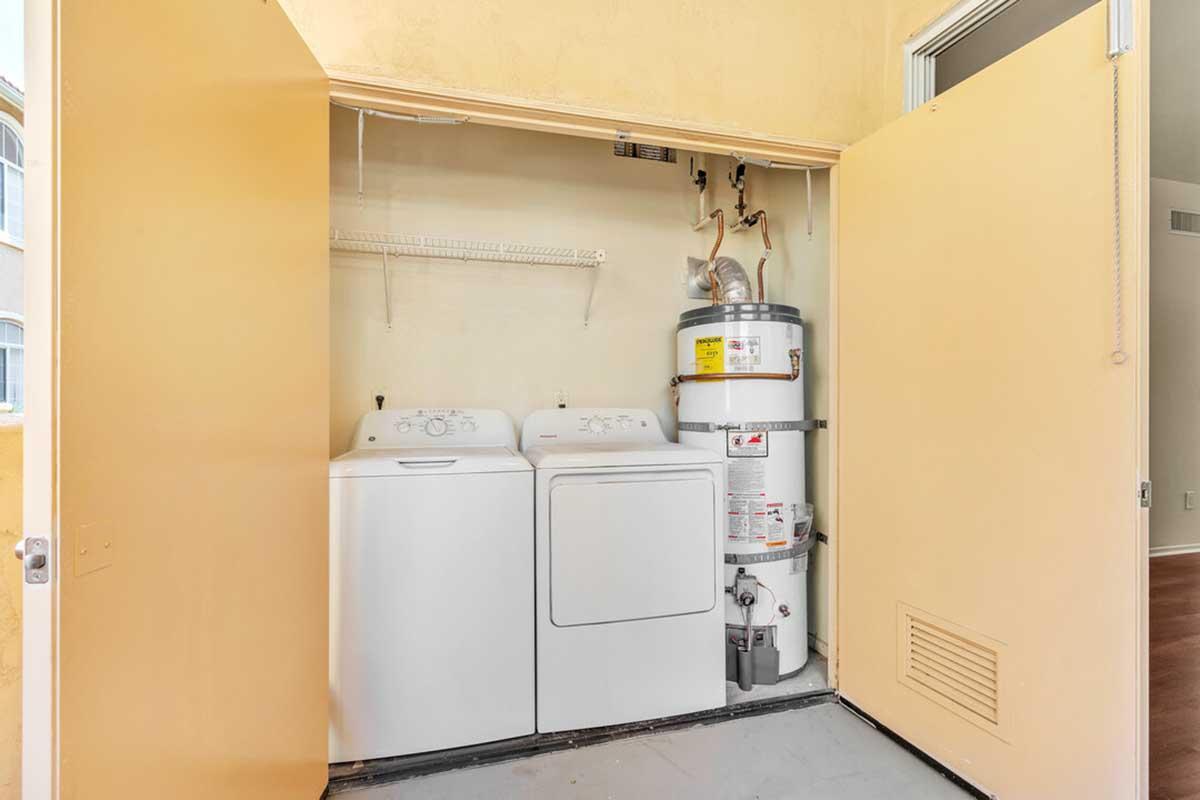 A laundry area featuring a white washing machine, a white dryer, and a water heater, all neatly enclosed within yellow cabinet doors. A shelf above provides storage, with parts of the wall and floor visible in the background.