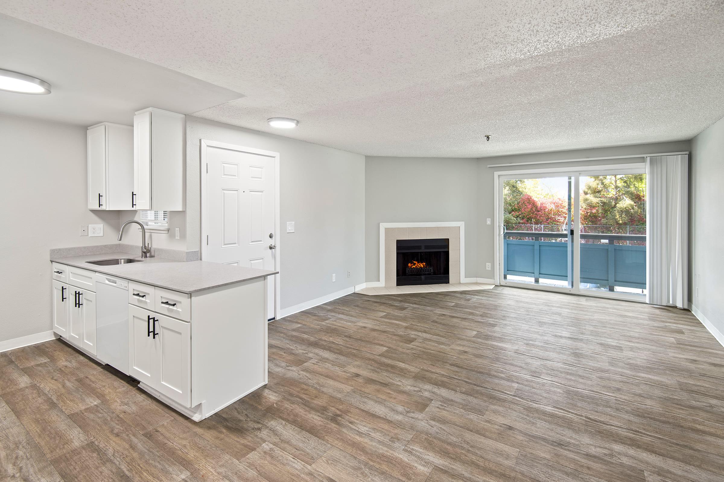 Open-concept living space featuring a modern kitchen with white cabinets and a granite countertop, a cozy fireplace, and large windows leading to a balcony. Neutral-colored walls and wood-style flooring create a bright, inviting atmosphere. Ideal for comfortable living and entertaining.
