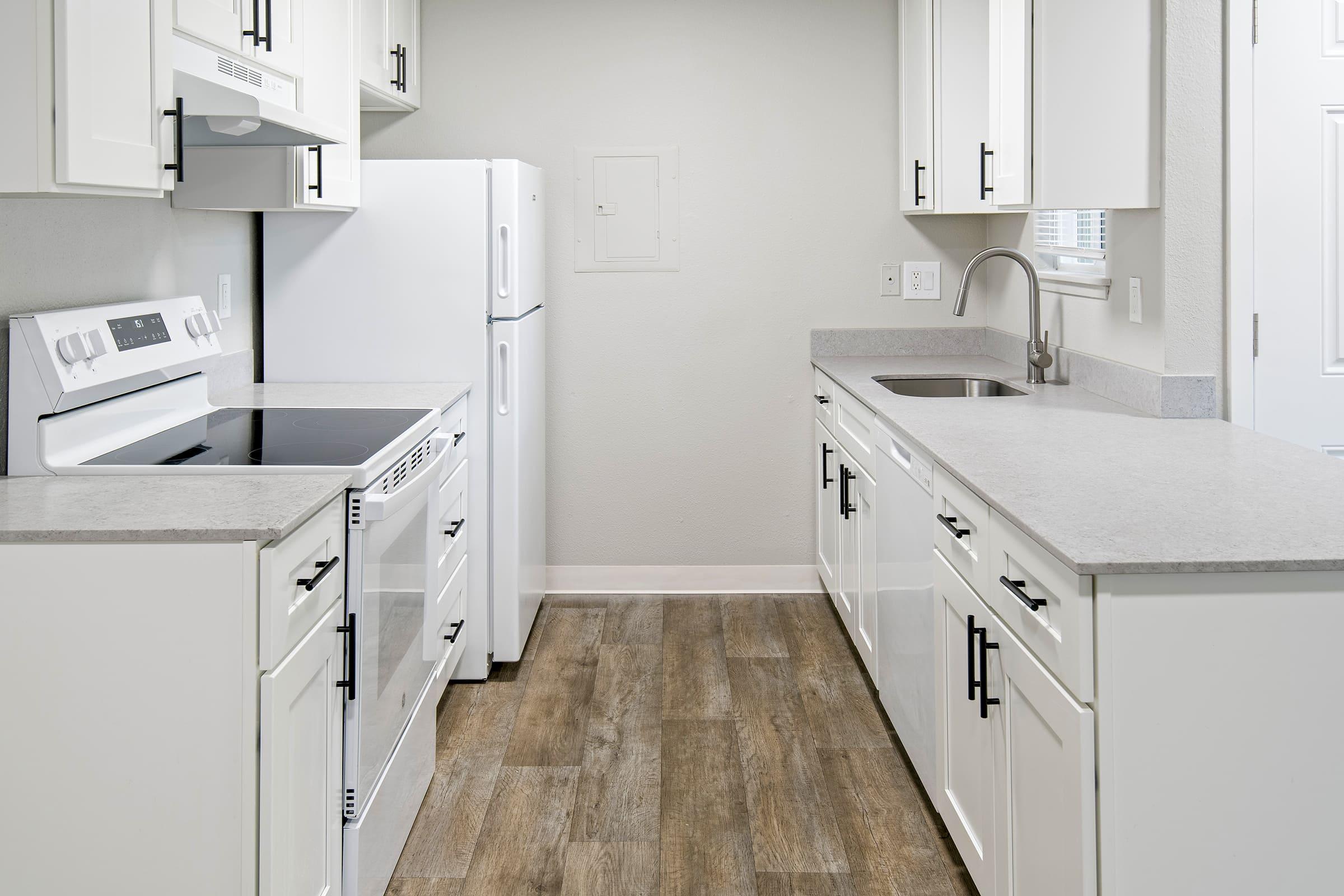 A modern kitchen with white cabinetry and gray countertops. Features include a stovetop, oven, refrigerator, and a sink. The flooring is a wood-like laminate, and the walls are painted in a light neutral color, creating a bright and open atmosphere.