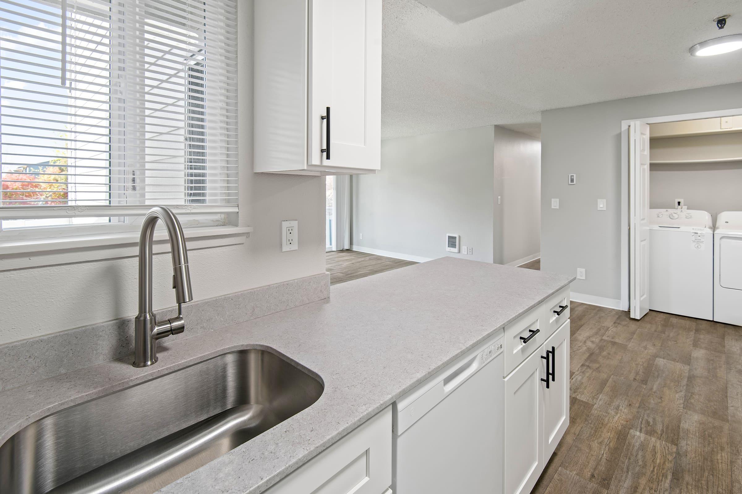 Modern kitchen with white cabinetry and a stainless steel sink. The countertop is light grey and there are large windows allowing natural light. In the background, a laundry area with washer and dryer is visible, and the room features neutral walls and wood-like flooring.
