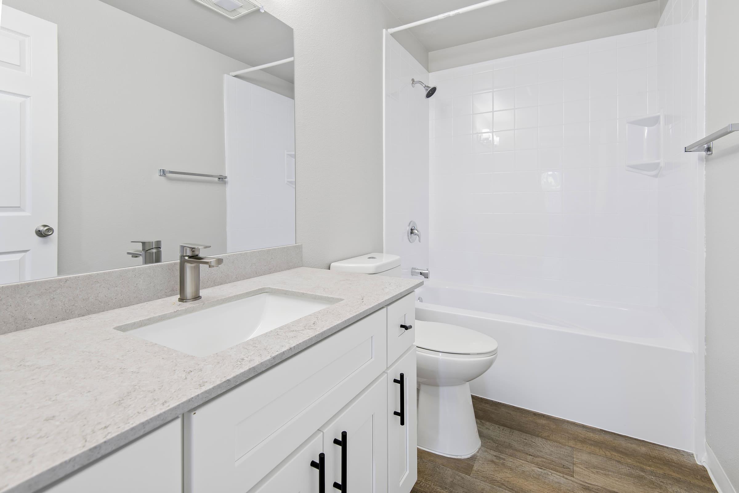 A clean and modern bathroom featuring a white bathtub, a white toilet, a light-colored countertop with a sink, and a mirror above the sink. The walls are painted in a soft tone, and there is a door visible on the left side. The overall design is bright and spacious.