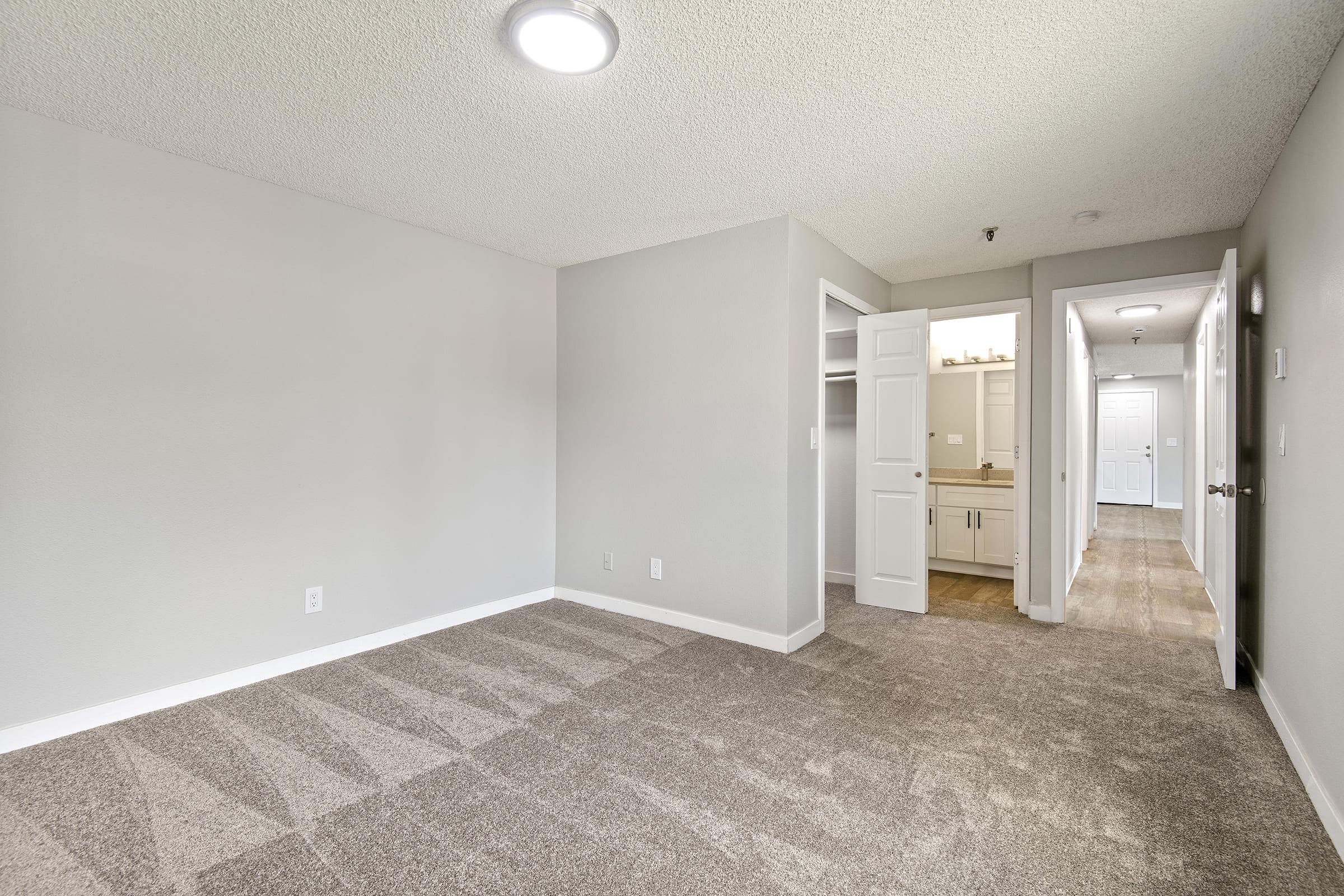 Empty room with light gray walls and plush carpeting. A closet is open on the left side, revealing shelving. In the background, a doorway leads to a hallway with additional interior doors. The ceiling features a circular light fixture, providing bright illumination.