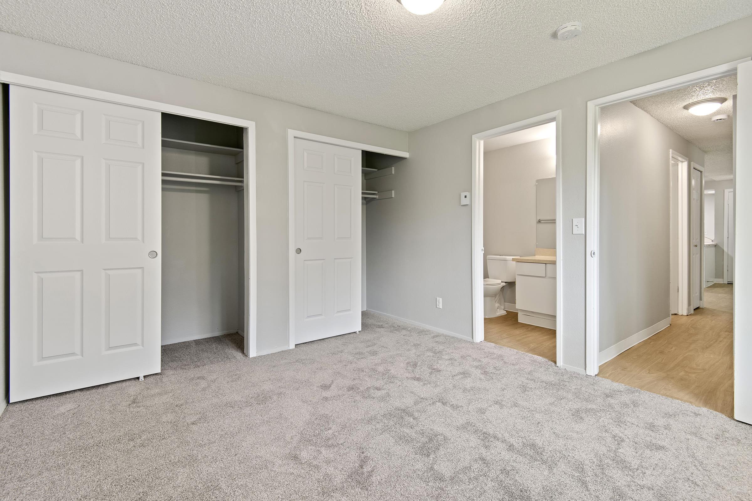 A spacious room featuring light gray walls and plush carpet. Two closet doors are visible on the left, while a doorway at the end leads to a bathroom. The room is well-lit, with a ceiling fixture providing additional illumination. Soft natural light may be entering from unseen windows.