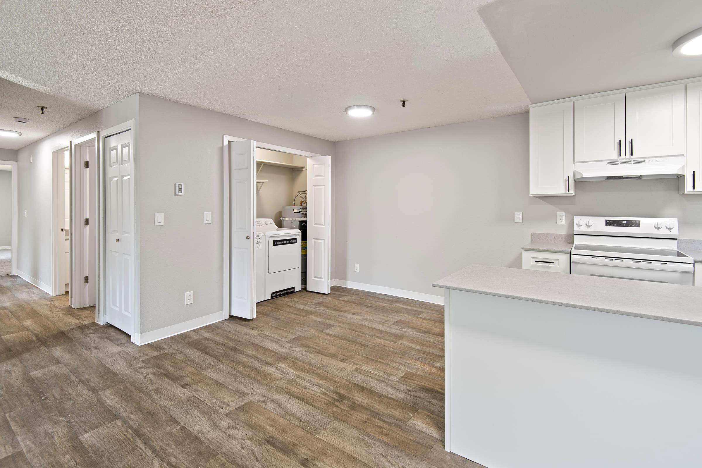 Bright, modern interior of an apartment featuring an open layout. The kitchen is equipped with white cabinetry and stainless steel appliances. A laundry area is visible in a separate room with doors open, and the flooring is a stylish wood-like laminate. Walls are painted in light neutral colors.